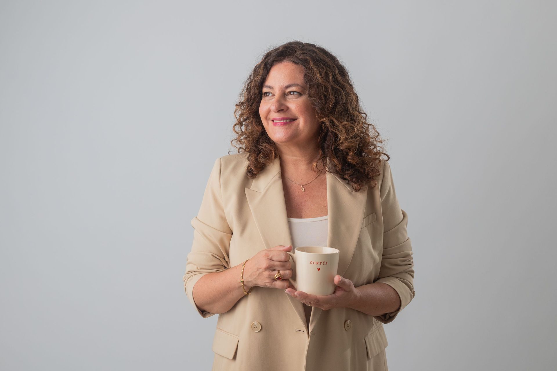Mujer con blazer beige sonriendo y sosteniendo una taza blanca sobre un fondo gris claro.