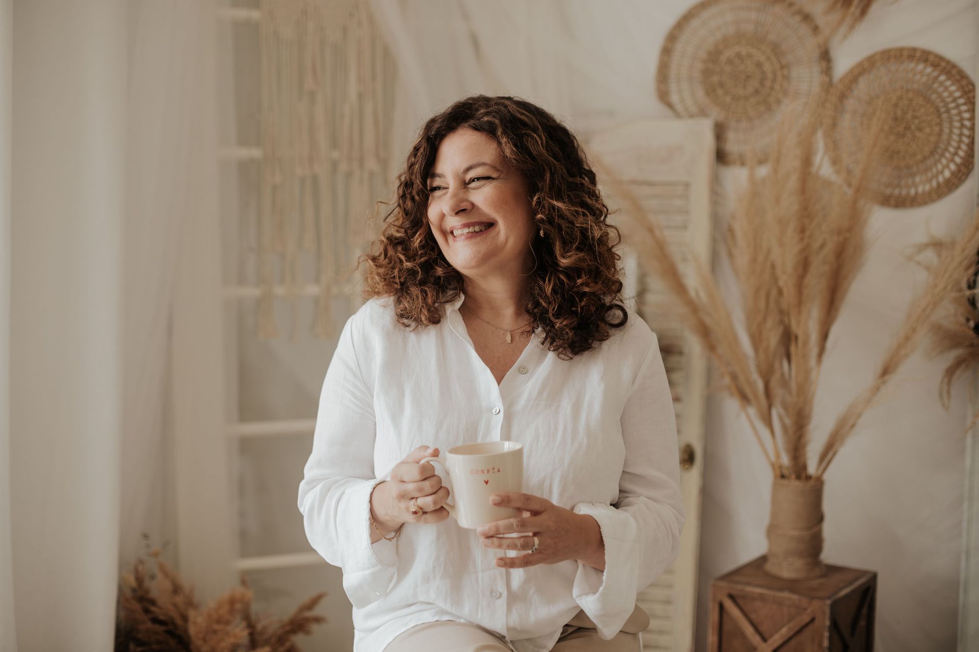 Mujer vestida de blanco sonriendo mientras sostiene una taza en una cálida habitación beige decorada con flores de la Pampa secas.