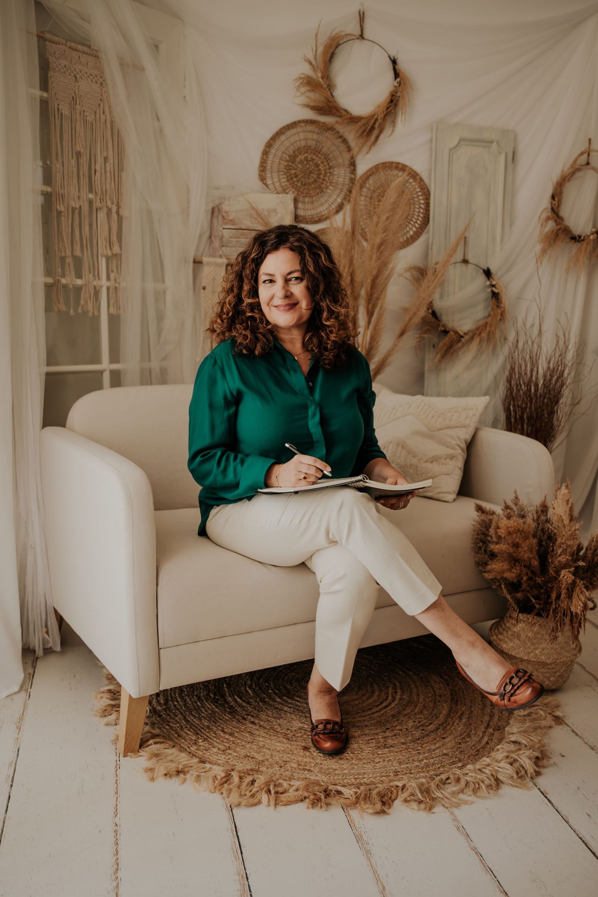 Una mujer sentada en una silla color crema en una acogedora habitación de estilo bohemio, sosteniendo un cuaderno y sonriendo.