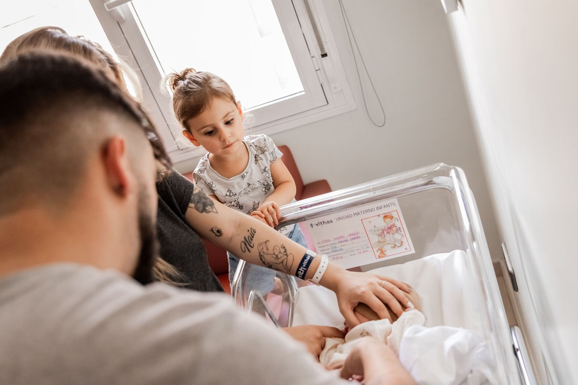 Un adulto consuela a un niño junto a una cuna de hospital cerca de una ventana luminosa.
