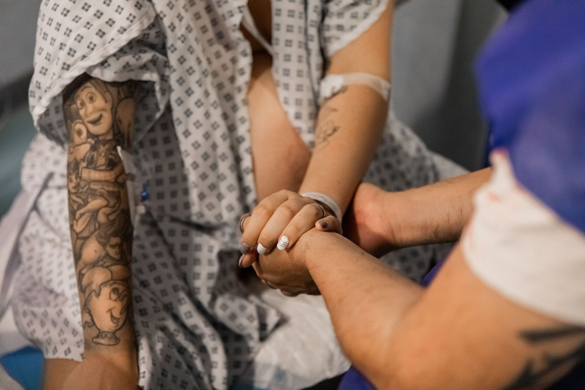 Manos entrelazadas en señal de apoyo, con un brazo tatuado y una bata de hospital visibles en un entorno clínico.