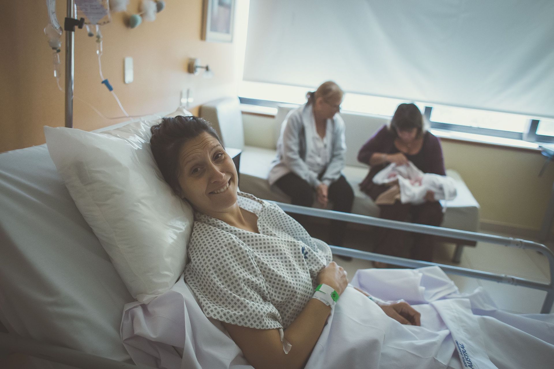 Paciente hospitalizada acostada en la cama con visitantes sentados a su lado cerca de una ventana luminosa.