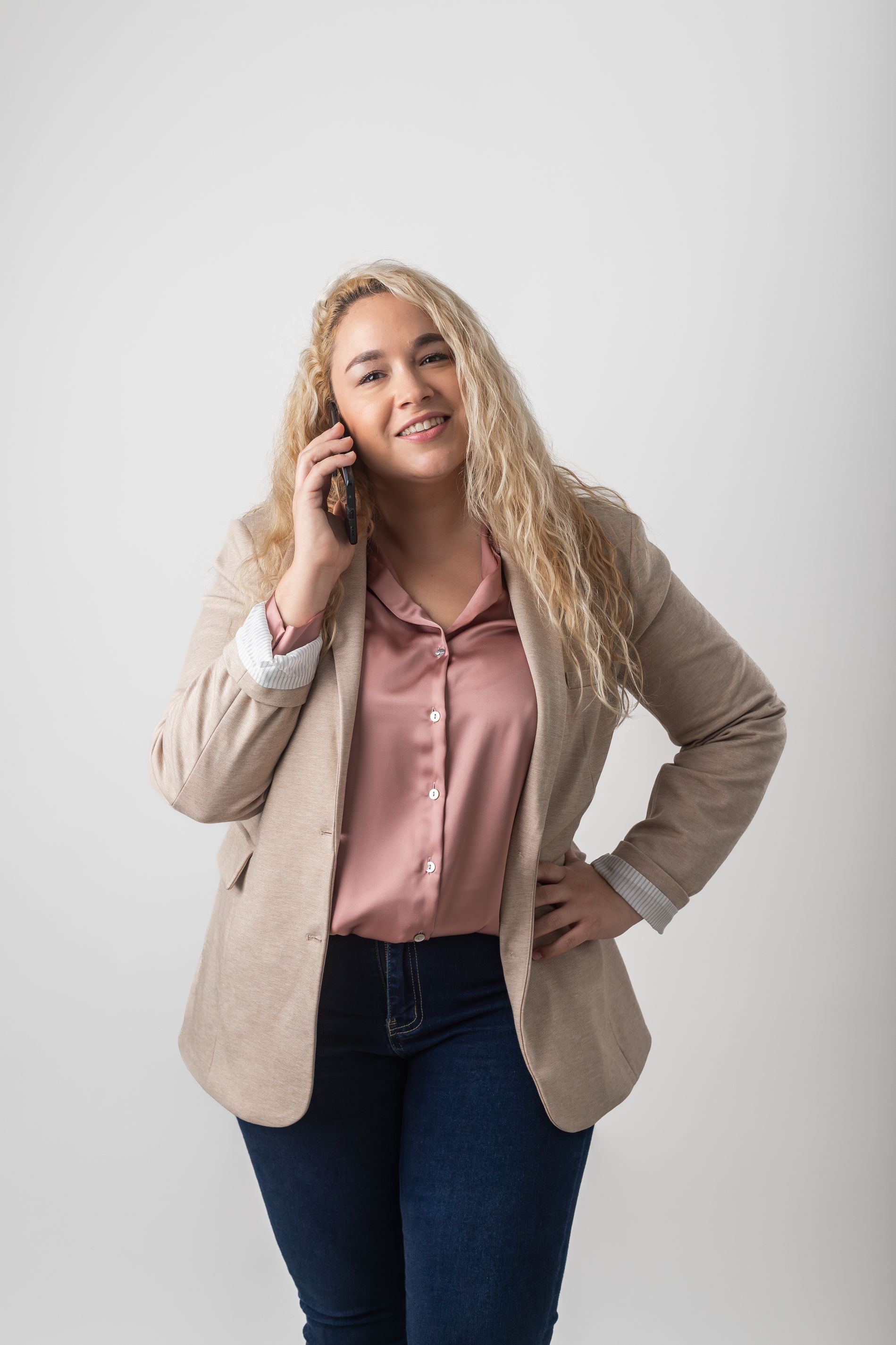 Mujer con blazer beige y blusa rosa sonriendo y sosteniendo un teléfono contra un fondo claro liso.