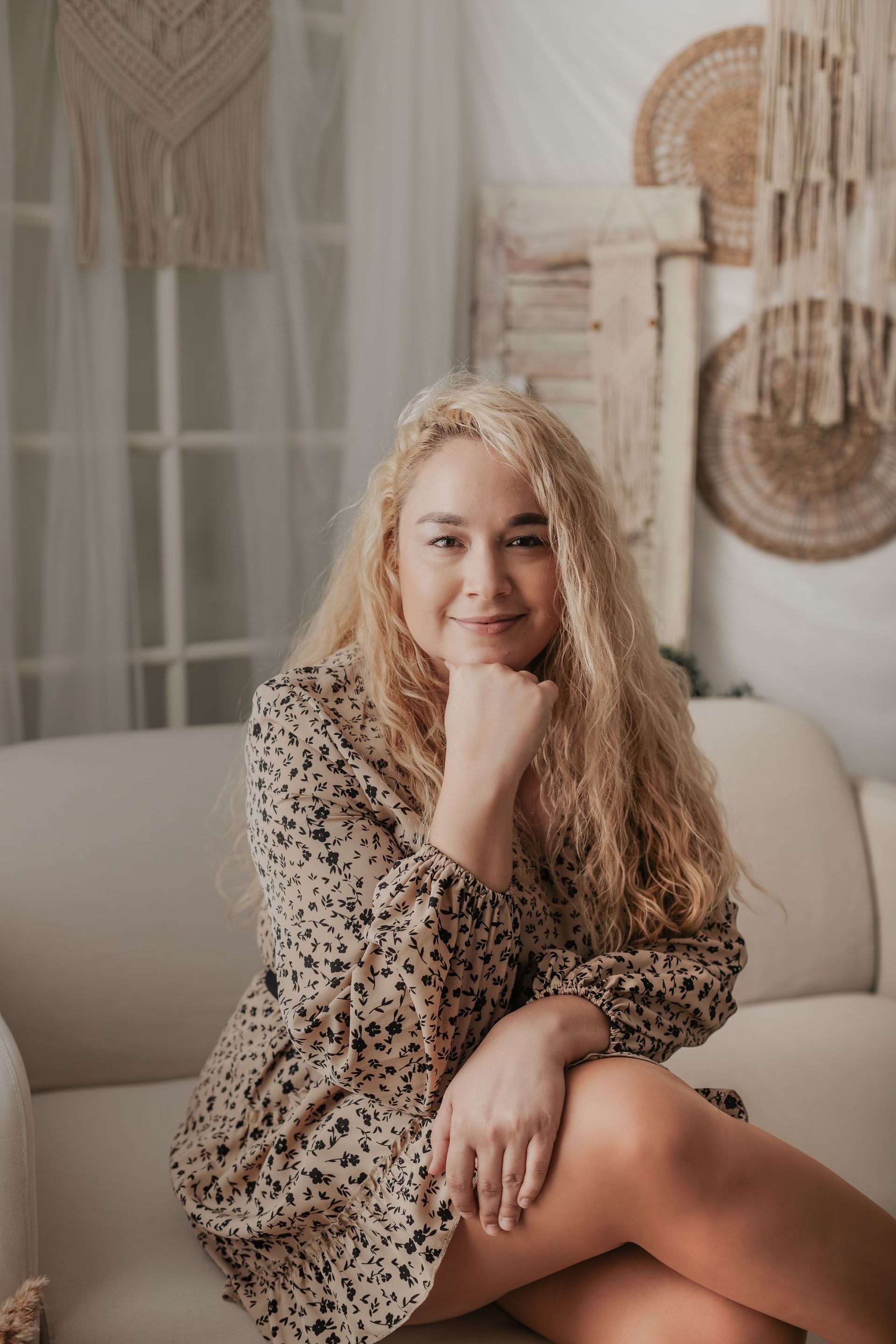 Una mujer sentada en un sofá beige en una habitación luminosa de estilo bohemio, sonríe con la barbilla apoyada en la mano.