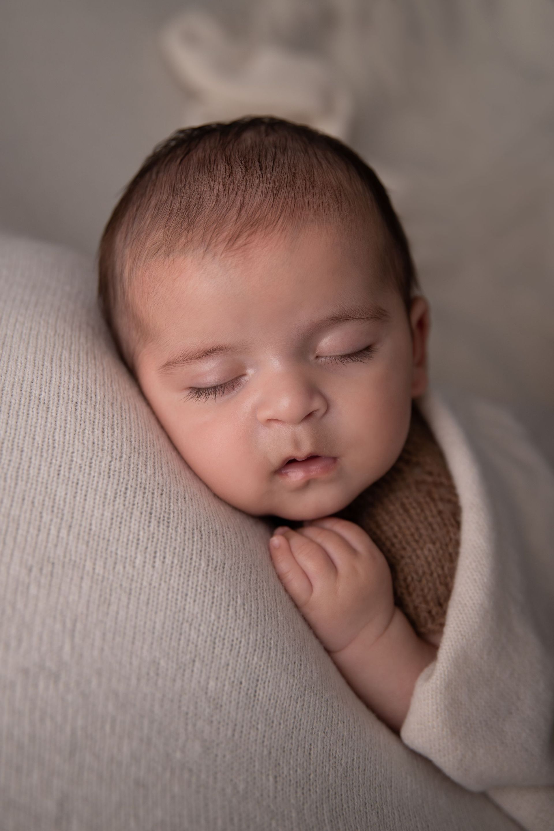 Un bebé dormido, envuelto en una suave manta blanca, descansa sobre un cojín beige con las manos entrelazadas.