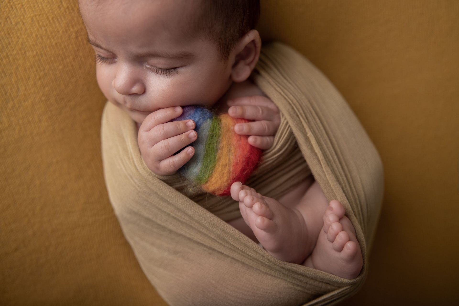 Bebé recién nacido envuelto en una manta beige, sosteniendo un corazón arcoíris del orgullo, descansando sobre una manta mostaza.