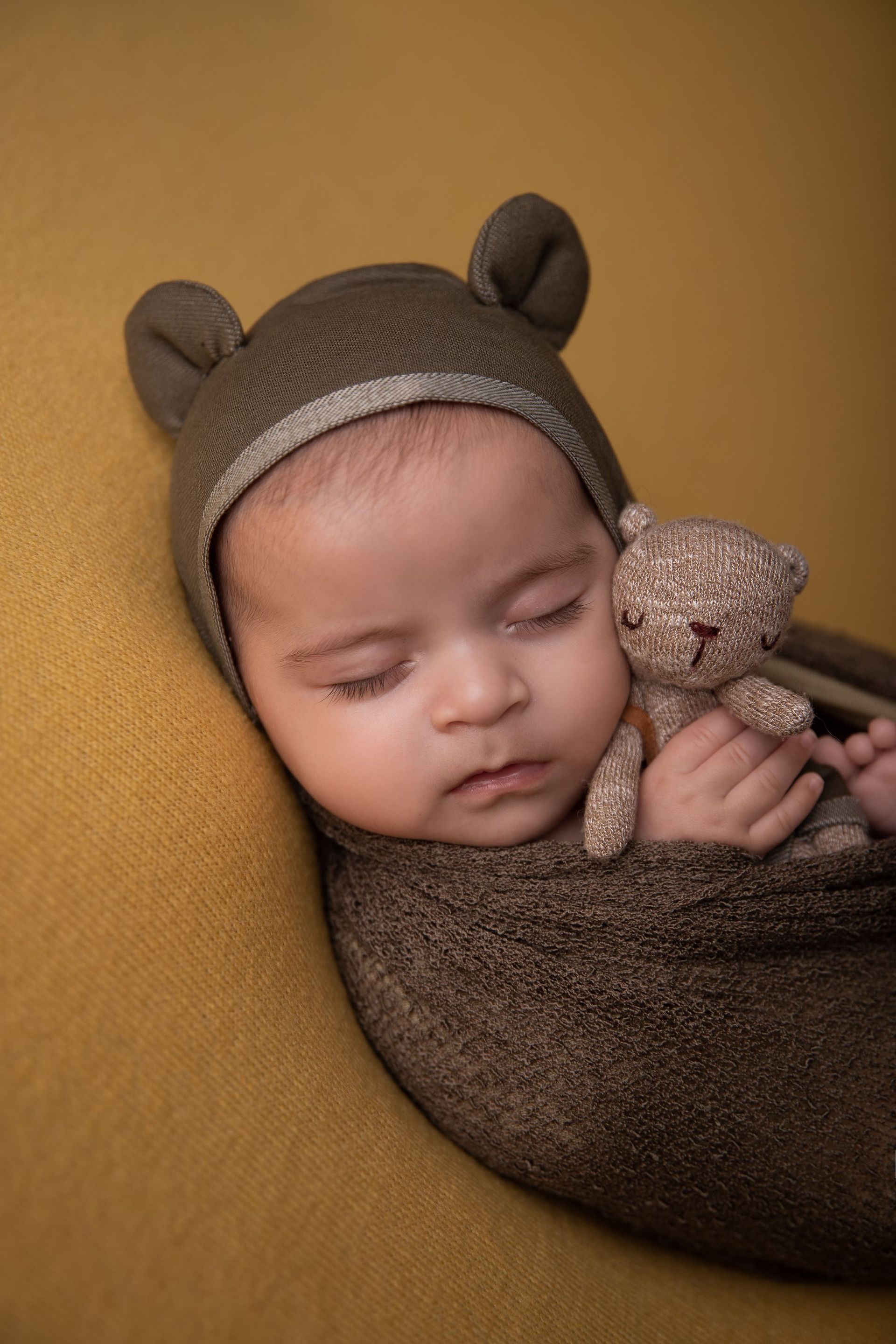 Un bebé dormido, envuelto en una manta marrón, con un gorro de oso y sosteniendo un pequeño peluche.