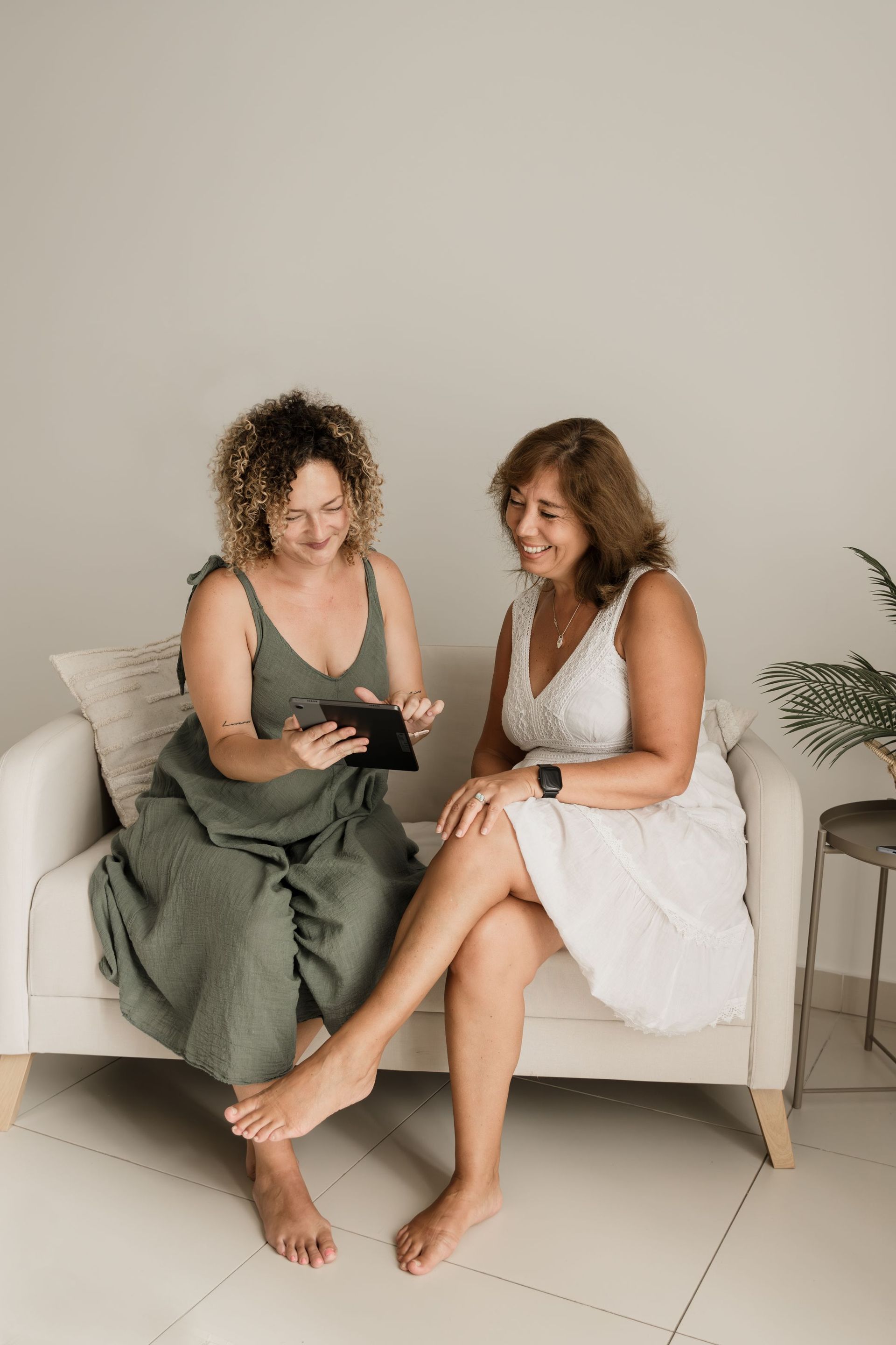 Dos mujeres están sentadas en un sofá, mirando una tableta en una sala de estar luminosa y minimalista.