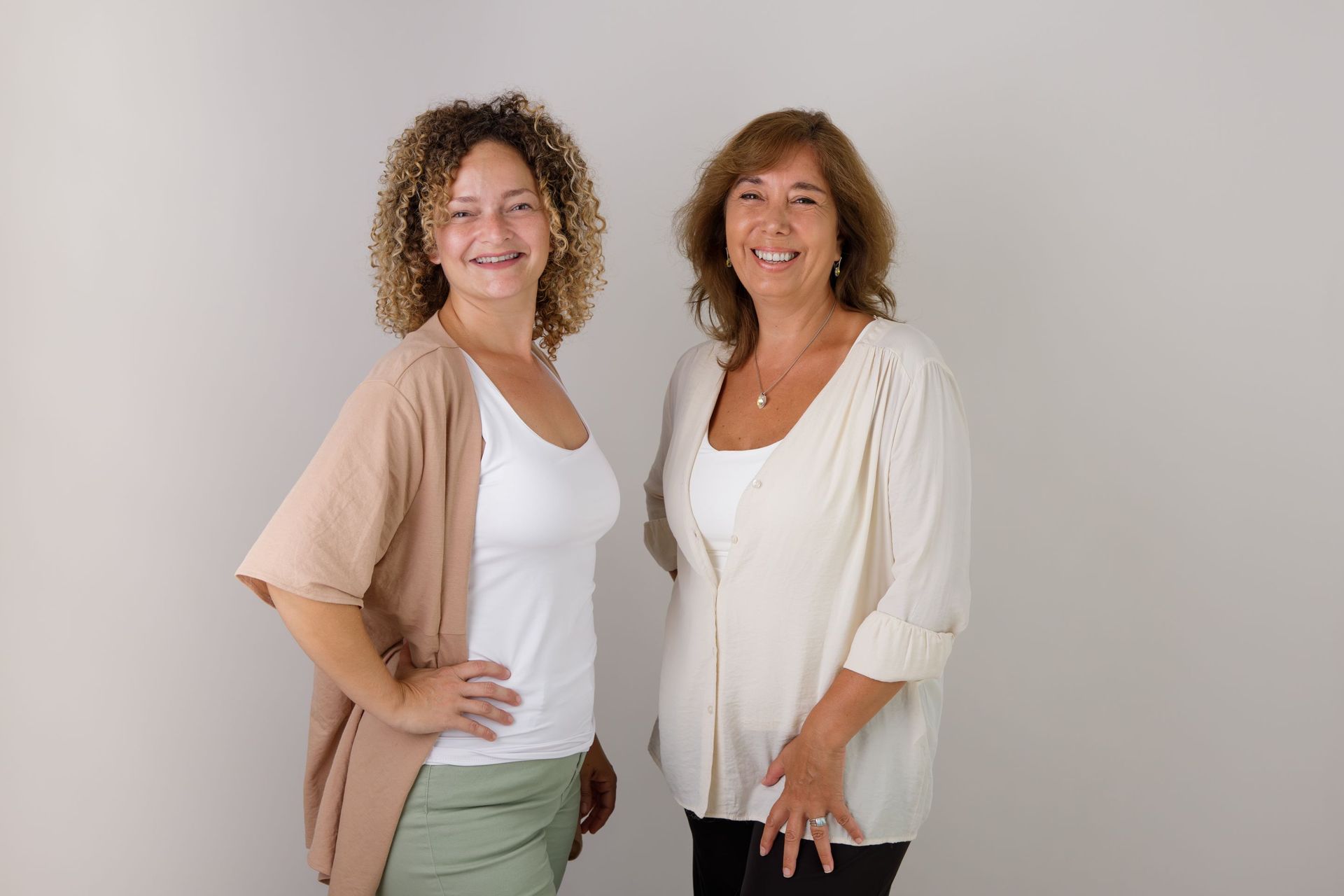 Dos mujeres sonrientes, de pie una al lado de la otra sobre un fondo liso, vestidas con ropa informal de colores claros.