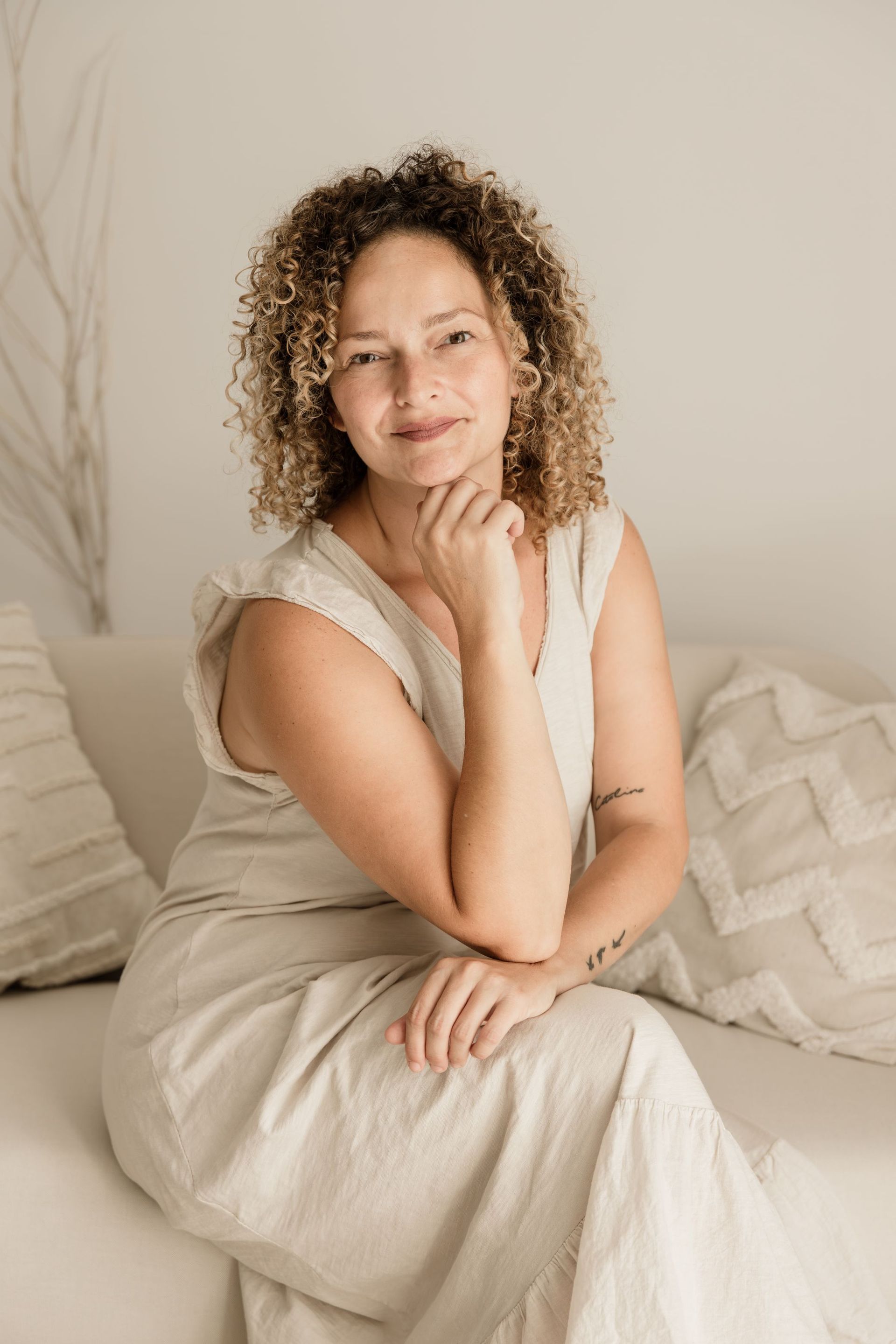 Mujer con un vestido color crema sentada en una cama, con la barbilla apoyada en la mano, en una habitación tenuemente iluminada.