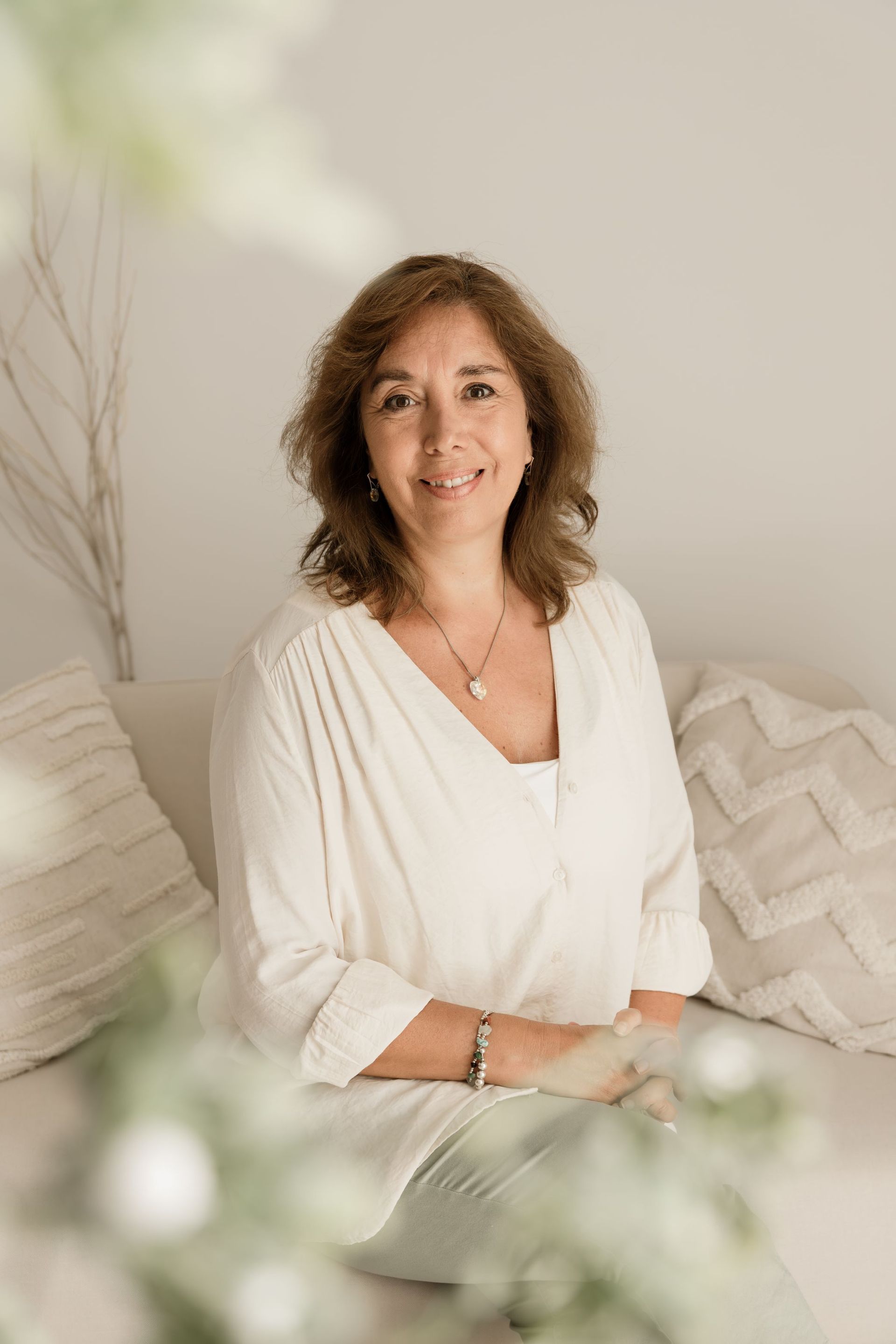 Mujer sentada en una cama en una habitación luminosa, con una blusa blanca y sonriendo dulcemente.