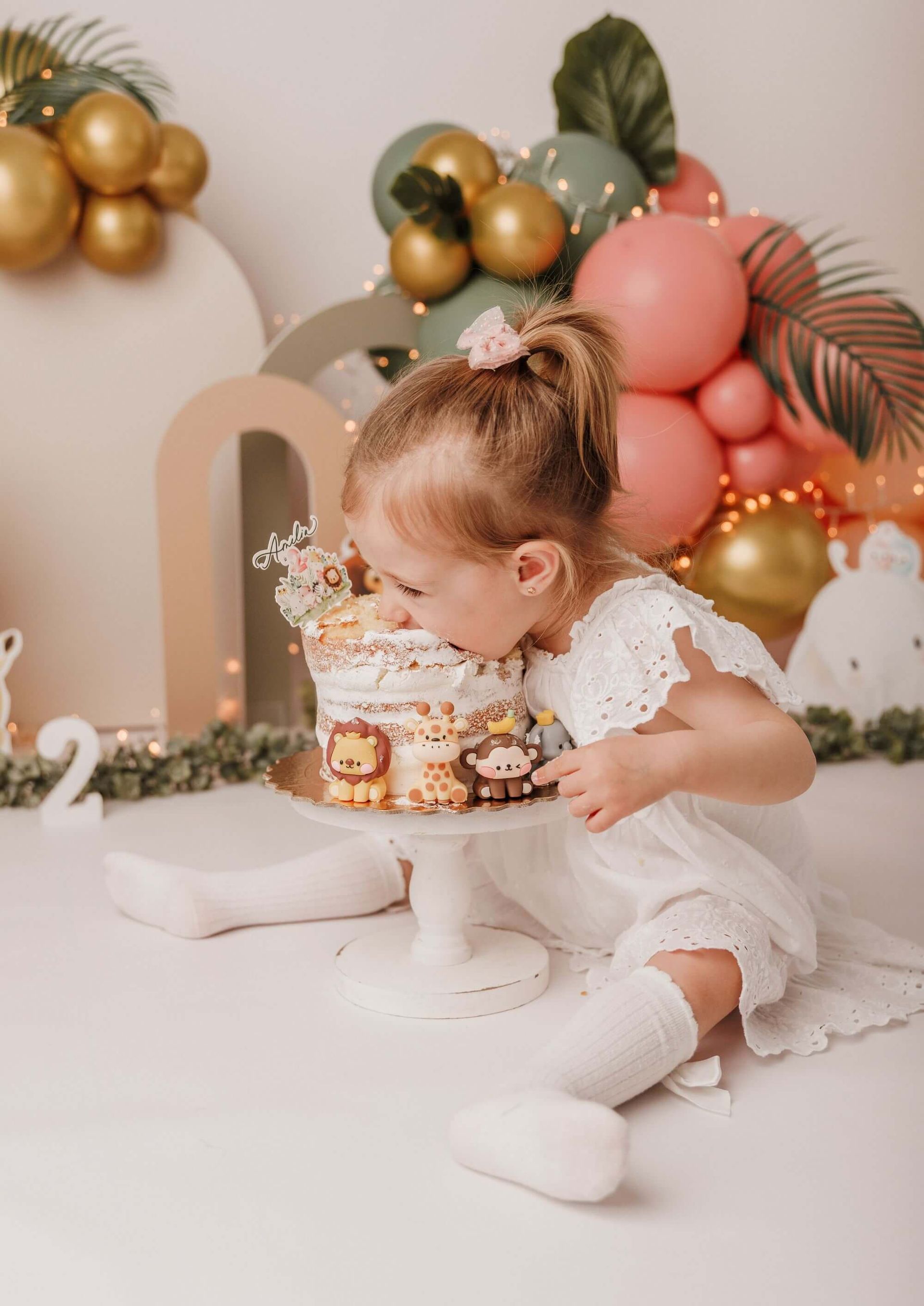 Una niña pequeña con un vestido blanco se inclina hacia un pastel en una fiesta de cumpleaños decorada con globos de colores pastel.