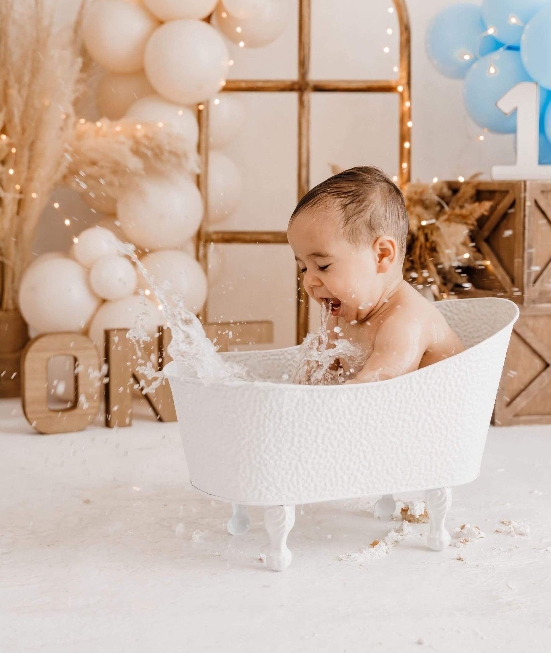 Un bebé chapoteando en una bañera blanca con un fondo de fiesta lleno de globos en tonos pastel.