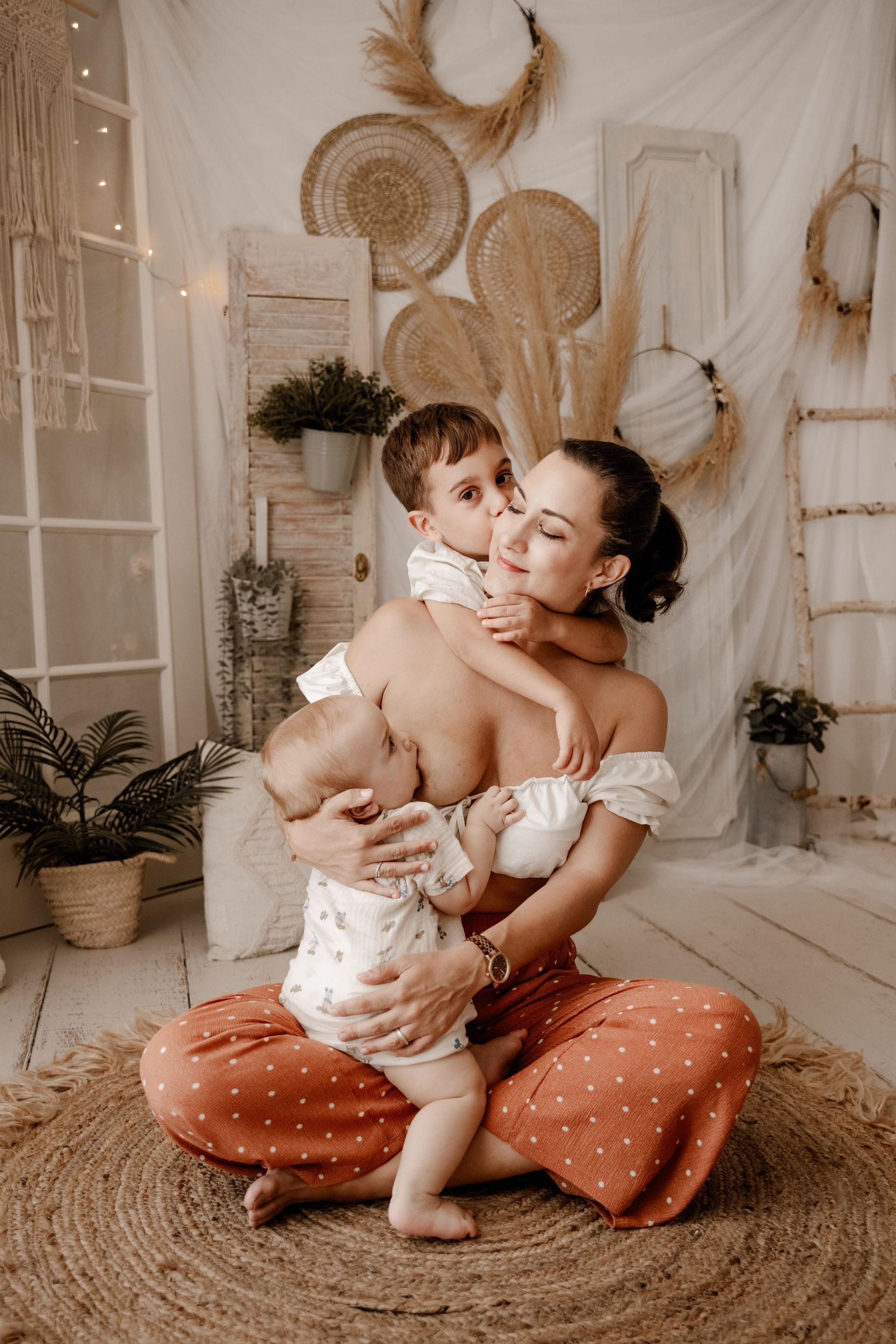 Una madre sentada en el suelo, sosteniendo a dos niños pequeños, en una acogedora habitación de estilo bohemio con decoración neutra.