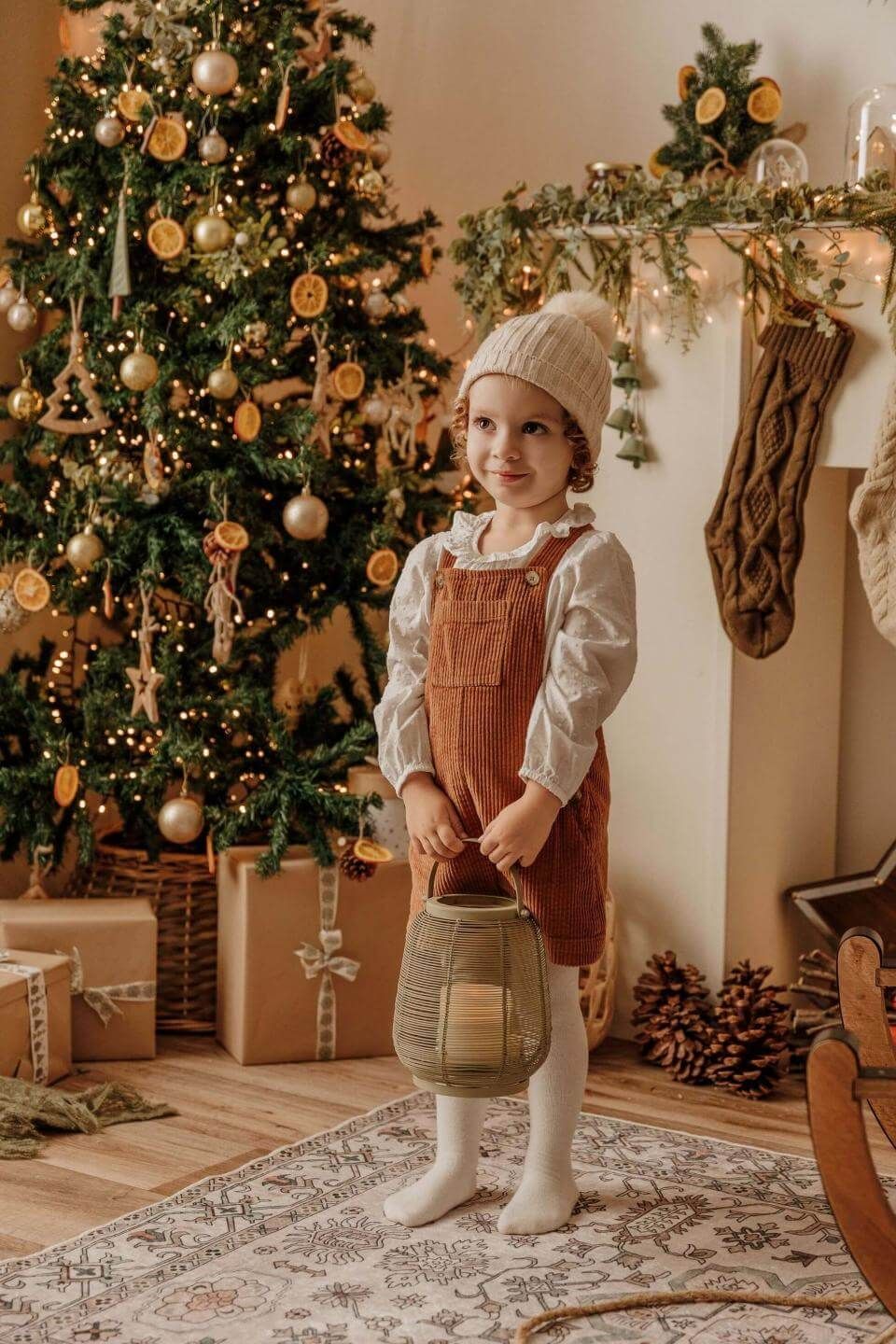 Niña con gorro de punto blanco y vestido color óxido de pie junto a un árbol de Navidad decorado