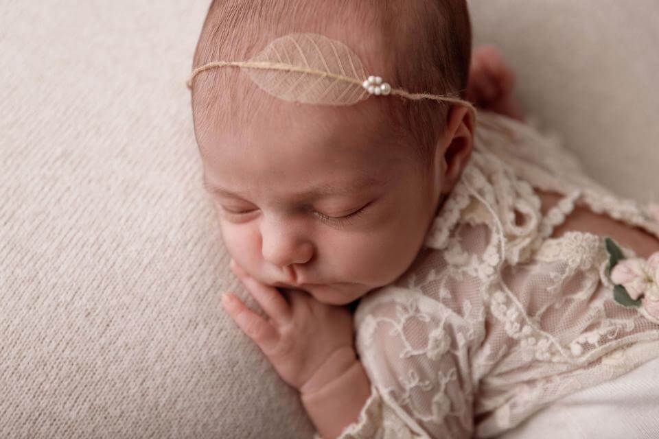 Bebé durmiendo con un conjunto de encaje color crema y una diadema, descansando sobre una suave manta beige.