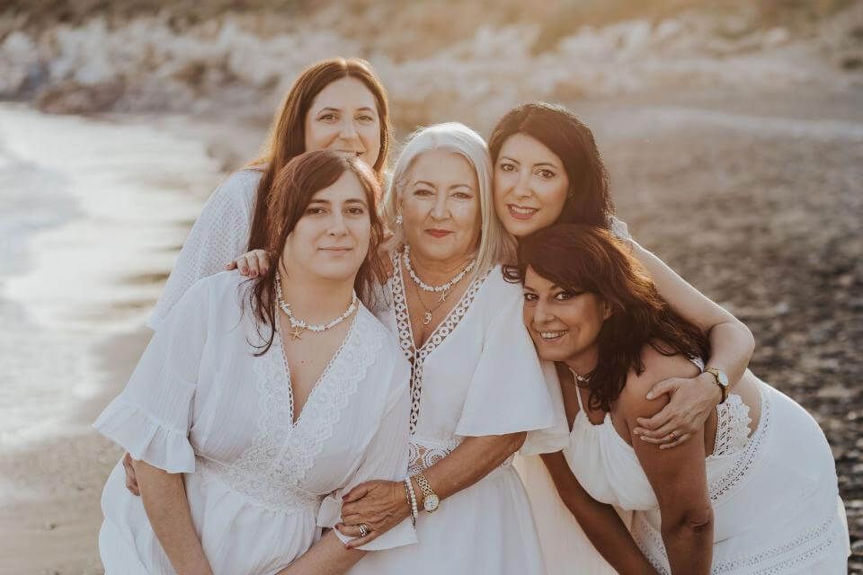 Cinco mujeres con vestidos blancos posando juntas en una playa al atardecer.