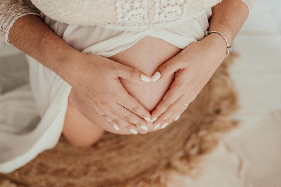 Manos apoyadas sobre el vientre de una mujer embarazada vestida con un vestido blanco sobre una alfombra suave.