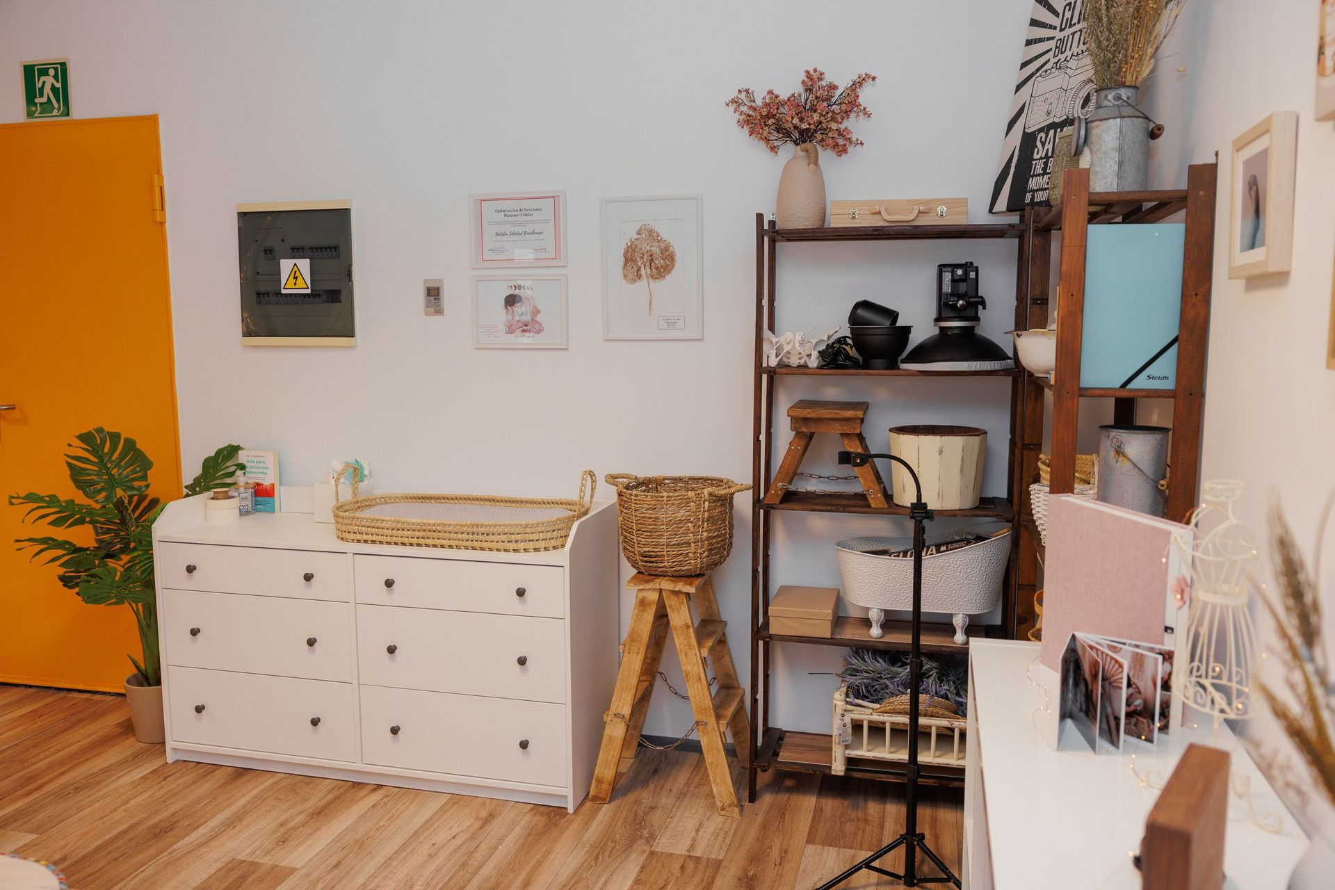 Acogedora sala de manualidades con cajones blancos, estante de madera, taburete de mimbre y plantas sobre suelo de madera.