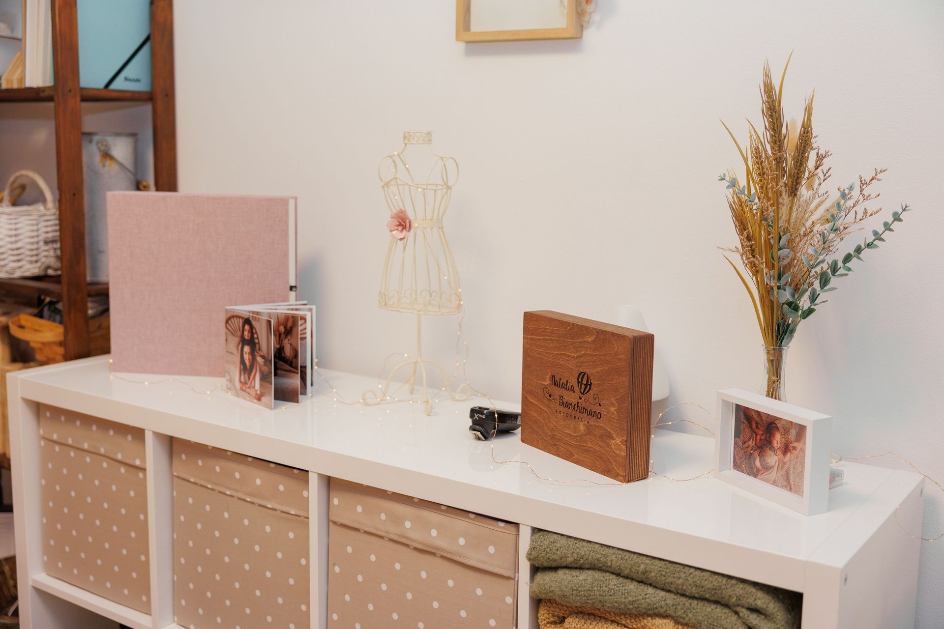 Mesa consola blanca con elementos decorativos, cuadros, libros y flores secas en una habitación luminosa.