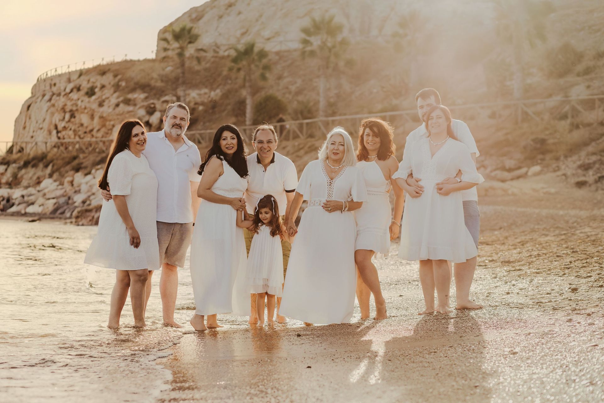 Grupo de personas vestidas de blanco de pie en una playa de arena al atardecer, sonriendo cerca de aguas poco profundas.