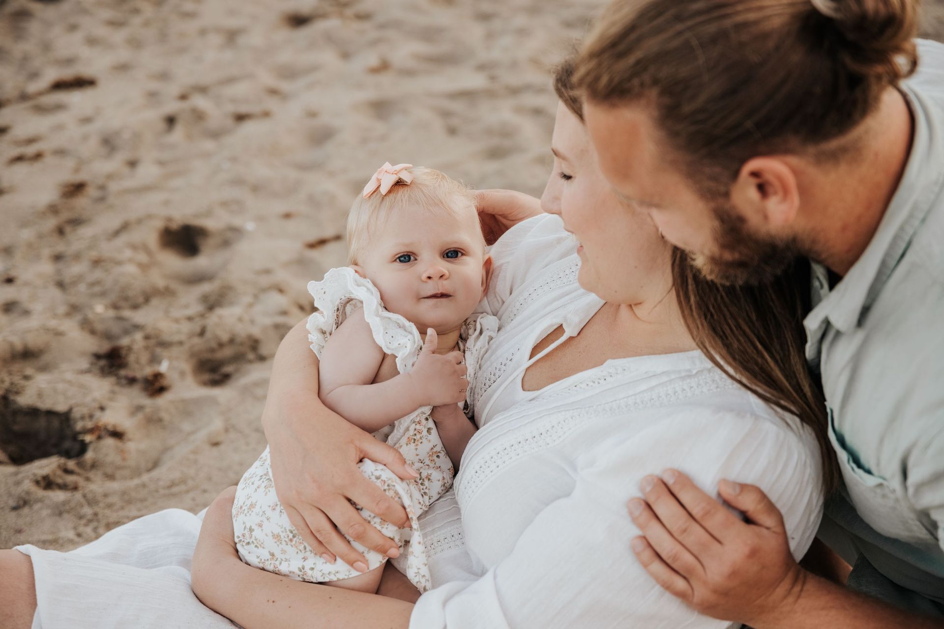 Un padre o una madre sostiene a su bebé en la playa, mientras otro adulto se inclina hacia él.