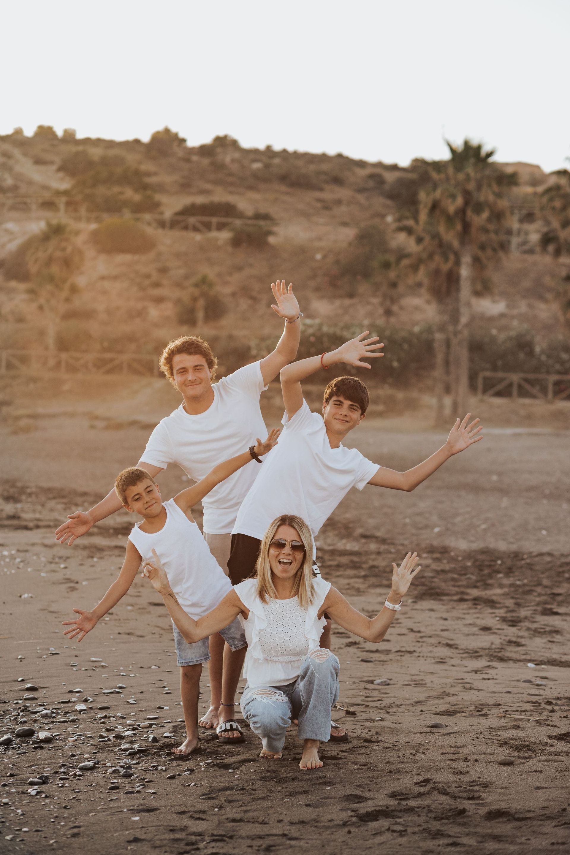 Cuatro personas posando y saludando en una playa de arena con colinas al fondo.