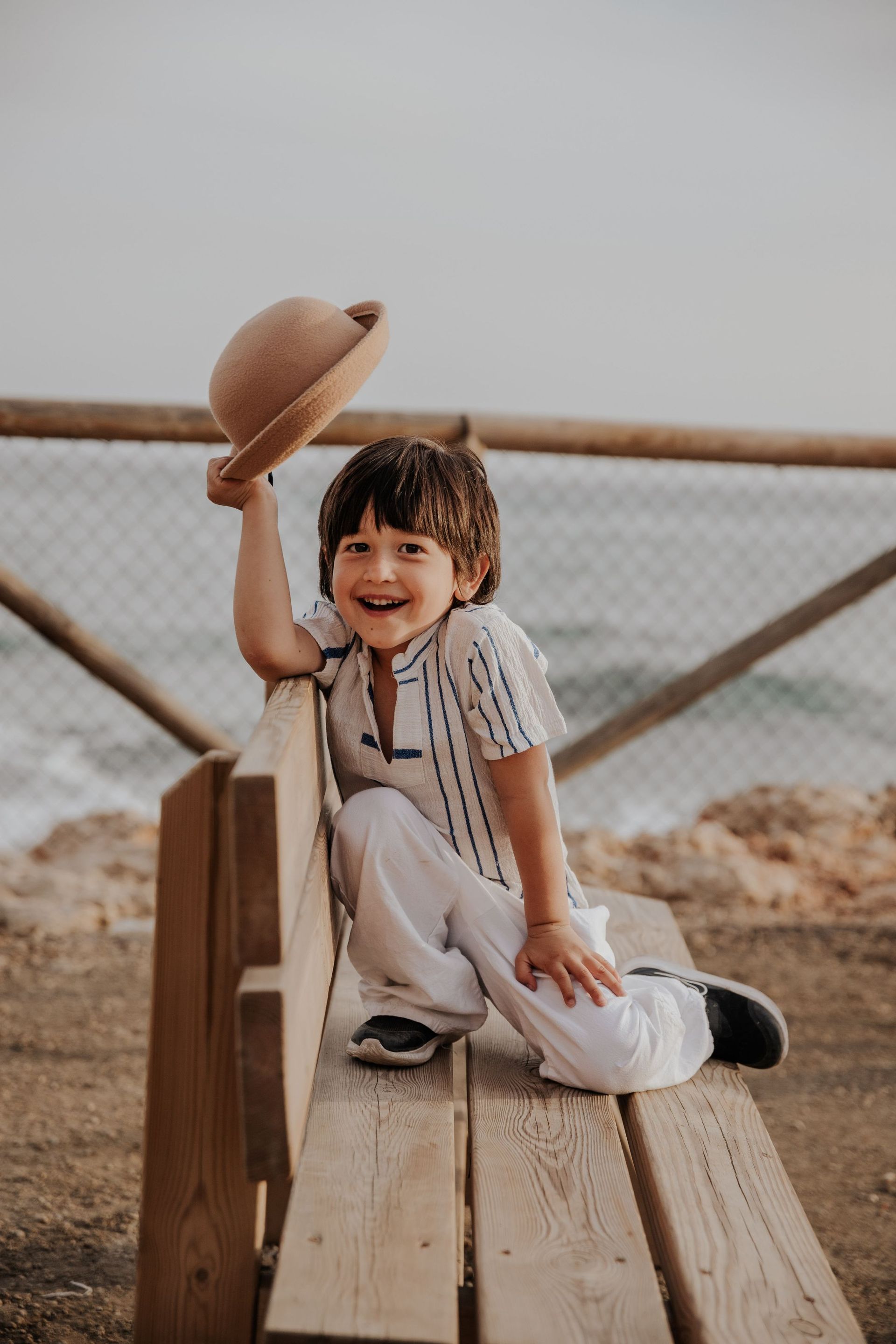 Un niño sonriente, vestido con una camisa de rayas y pantalones blancos, está sentado en un puente de madera