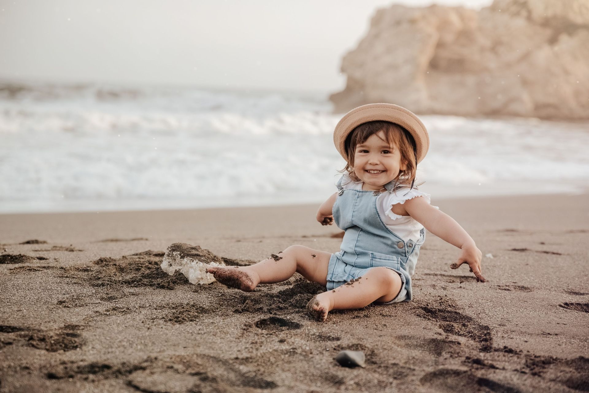 Un niño sonriente con un mono vaquero y un sombrero está sentado en una playa de arena junto al océano.
