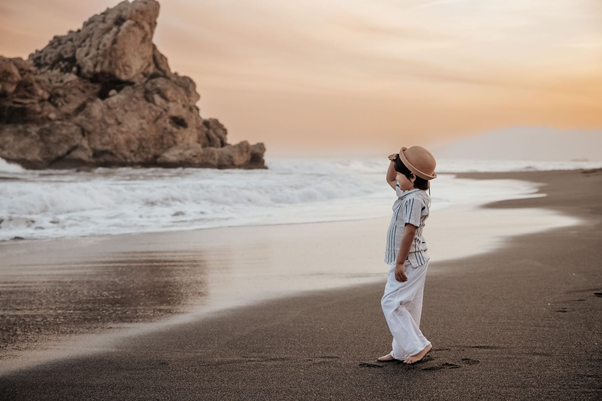 Una persona vestida de blanco y con sombrero de paja está de pie en una playa cerca de acantilados rocosos al atardecer.