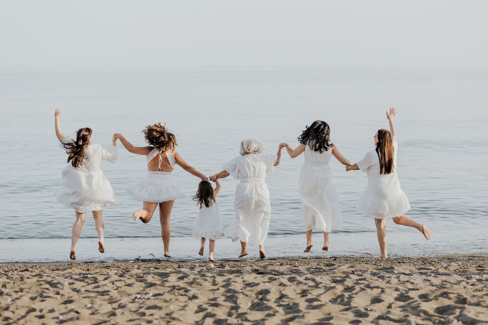 Cinco personas vestidas de blanco saltan de la mano en una playa frente al océano.