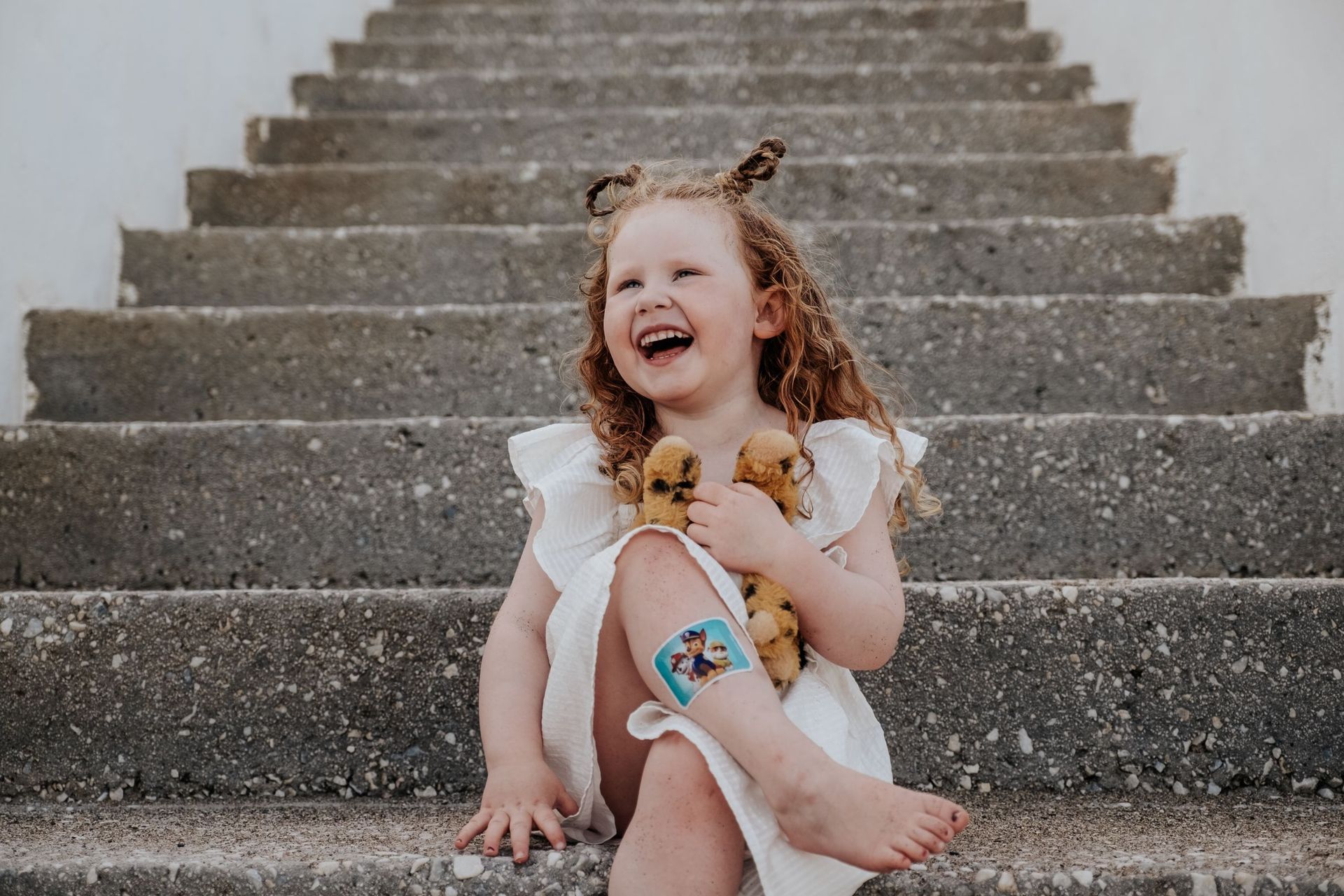 Niña sonriente con vestido blanco sentada en escalones de cemento con una flor en el pelo.