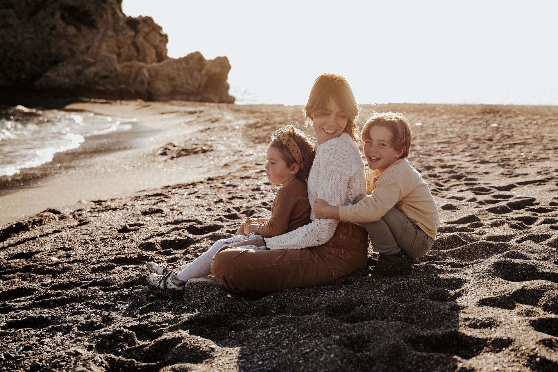 Tres niños sentados juntos en una playa de arena junto al agua al atardecer.