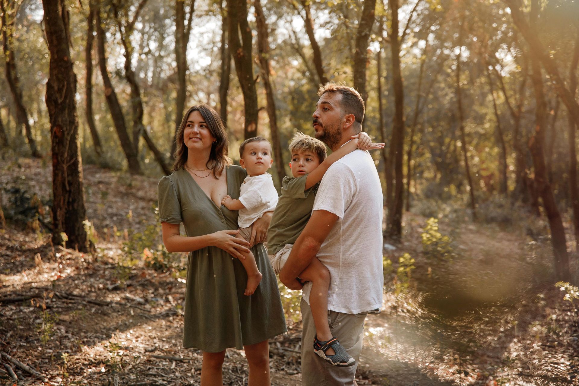 Una familia de cuatro personas de pie en un bosque iluminado por el sol, con un bebé y un niño en brazos