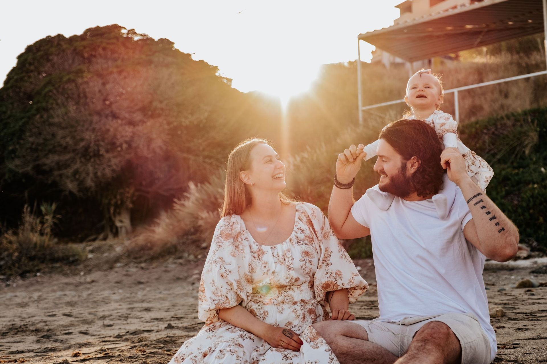 Una familia sentada en la playa al atardecer, sonriendo, con un niño sobre los hombros de un hombre.