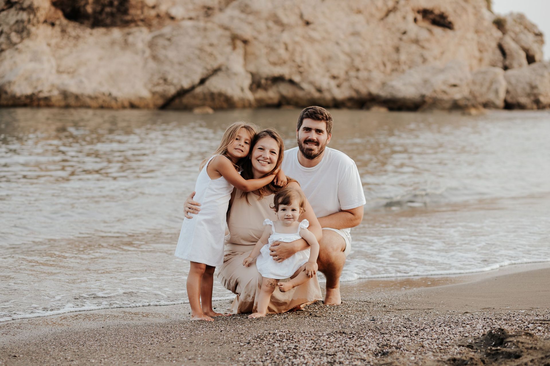 Familia de cuatro personas posando en una playa de guijarros, sonriendo y abrazándose cerca de la orilla.