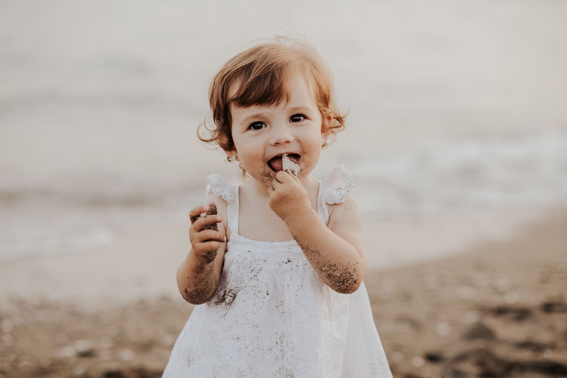 Una niña pequeña con un vestido blanco sonríe en la playa, sosteniendo y comiendo un pequeño refrigerio.