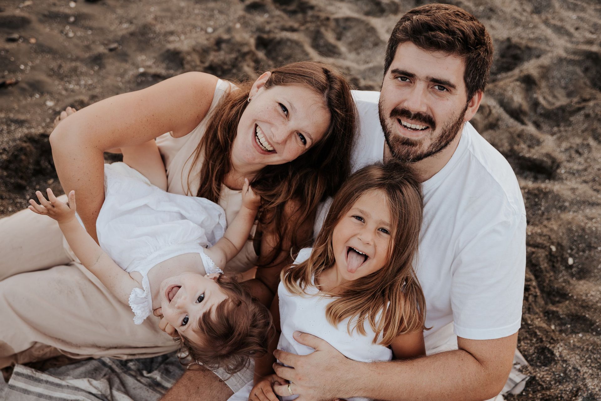 Una familia sonríe en una playa rocosa, apoyada en el suelo, vestida de blanco, con un perro delante.