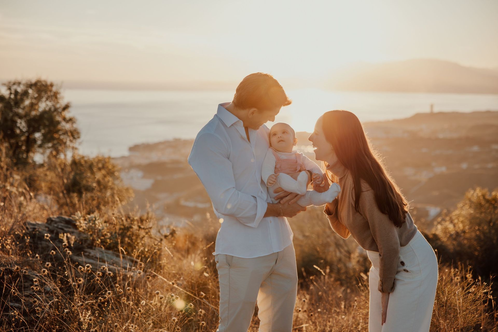 Familia al aire libre al atardecer, los padres sonríen a su bebé en un campo cálido y dorado.