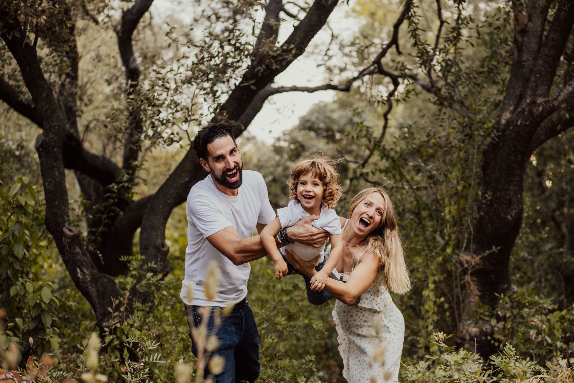 Familia sonriente tomada de la mano en un huerto arbolado, con un niño en brazos de dos adultos.