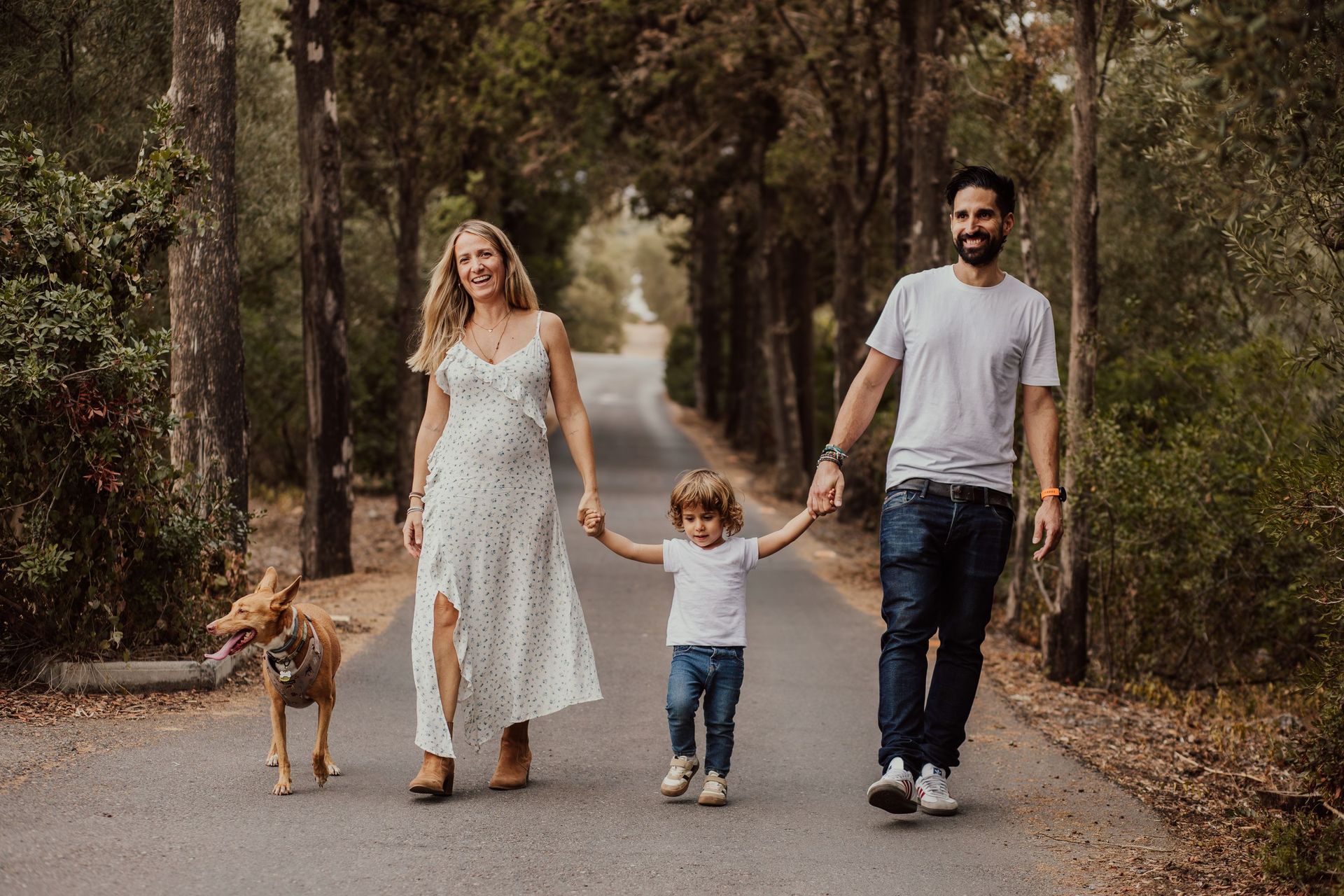 Una familia pasea con su perro por un camino arbolado, tomados de la mano y sonriendo.