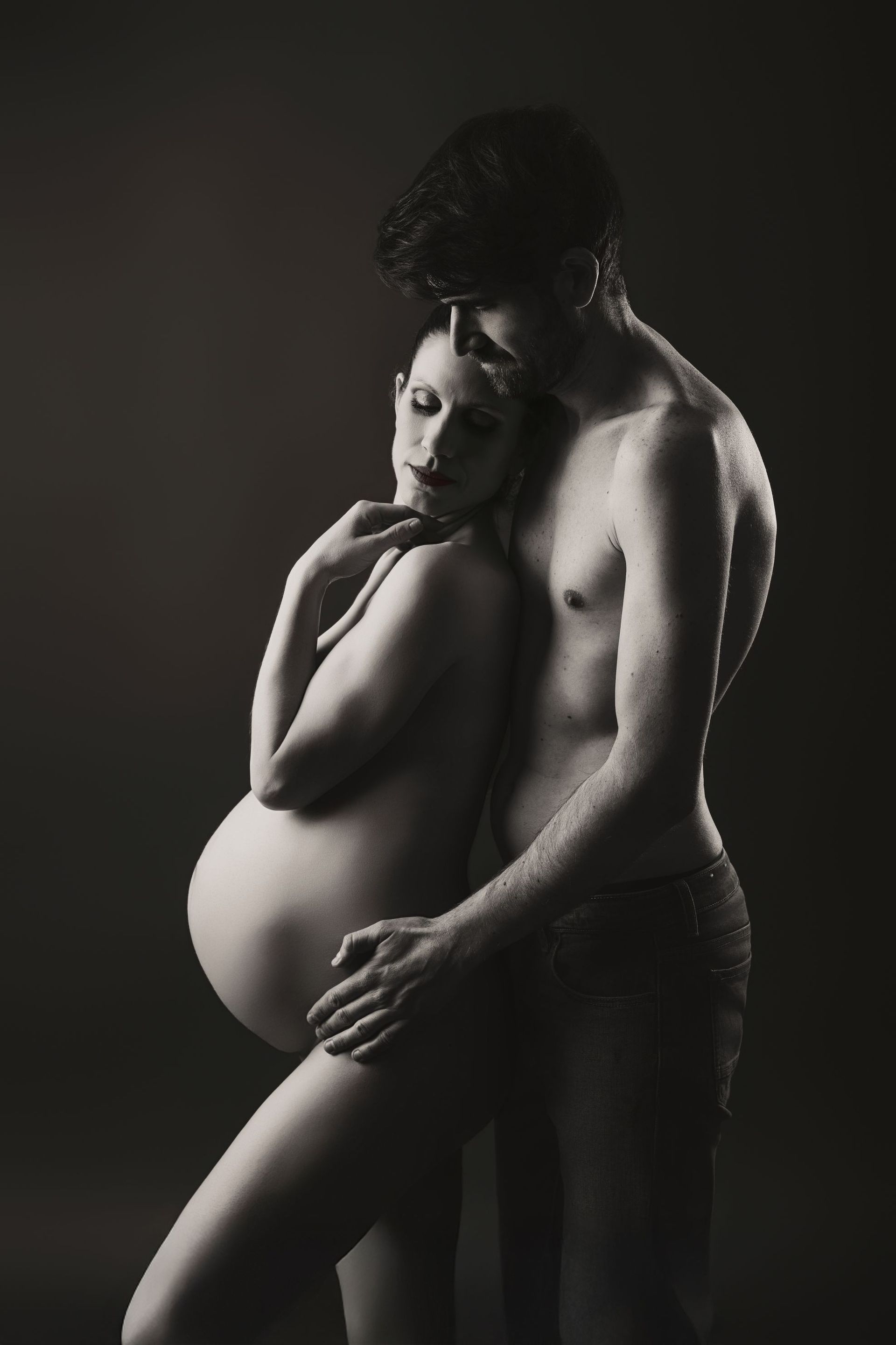 Mujer embarazada con su pareja, abrazándose en un retrato de estudio oscuro.