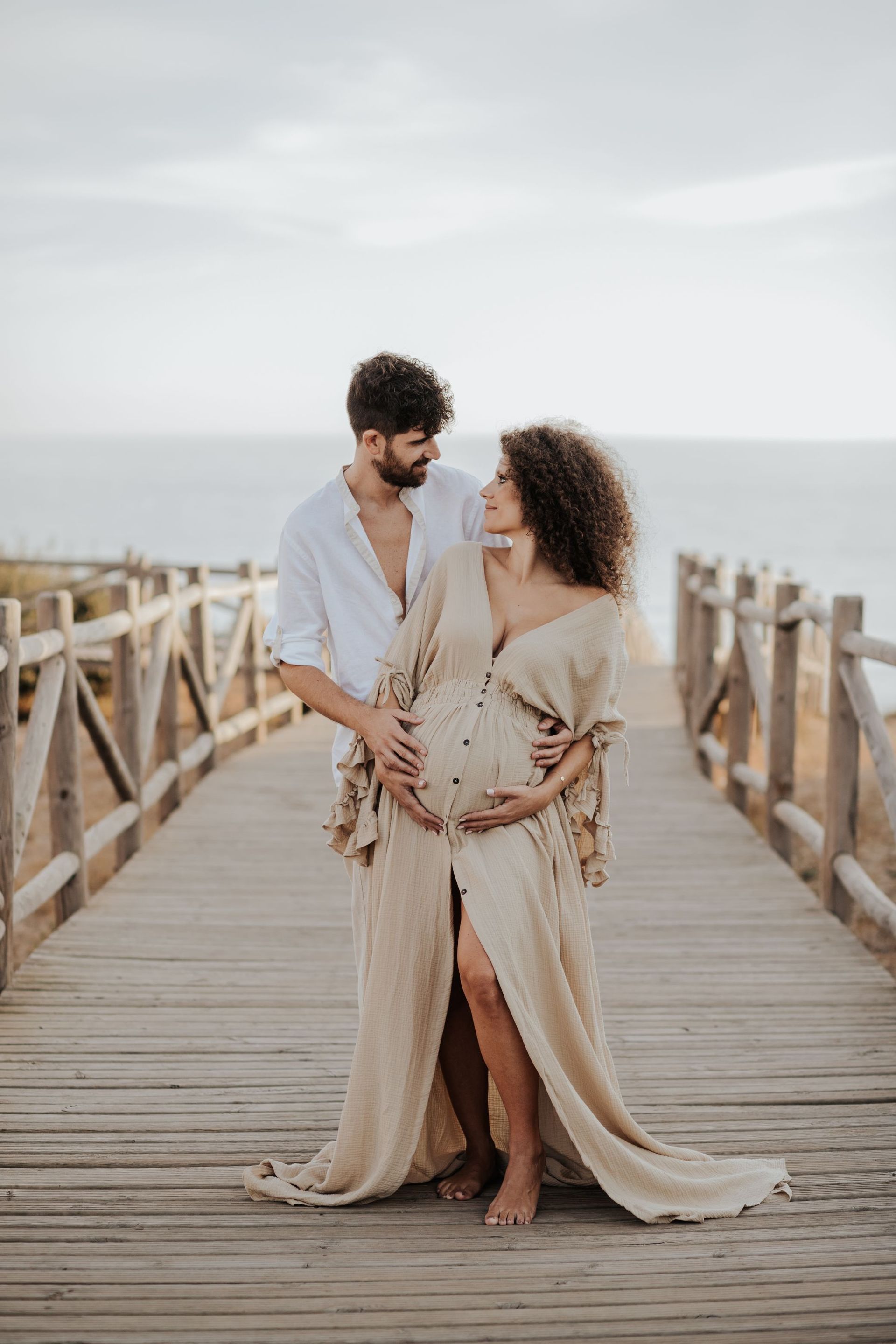 Una pareja posa en un muelle de madera junto al mar, compartiendo una escena íntima con ropa de playa ligera.