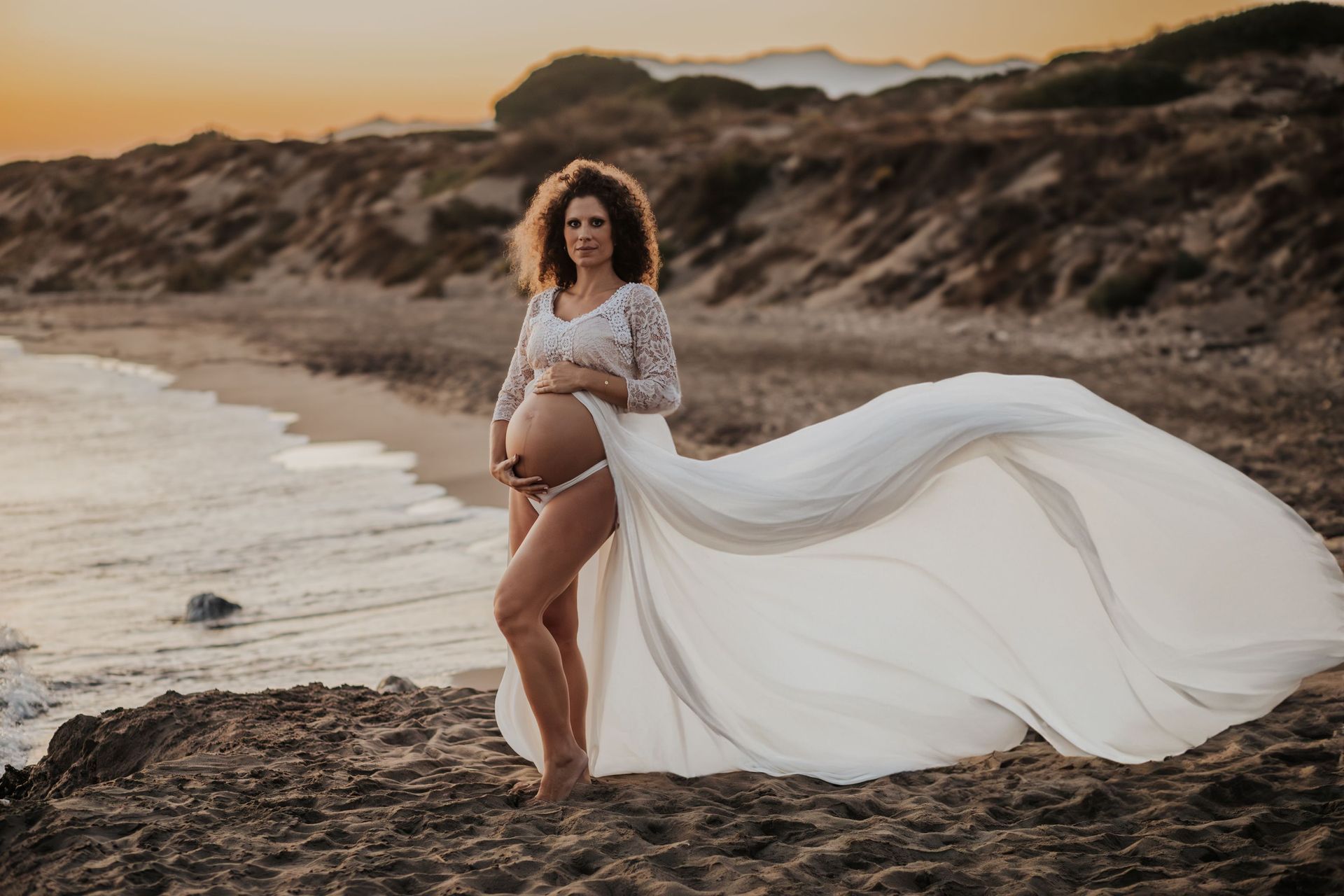 Mujer embarazada con un vestido blanco vaporoso de pie en una playa al atardecer, con la tela ondeando al viento.