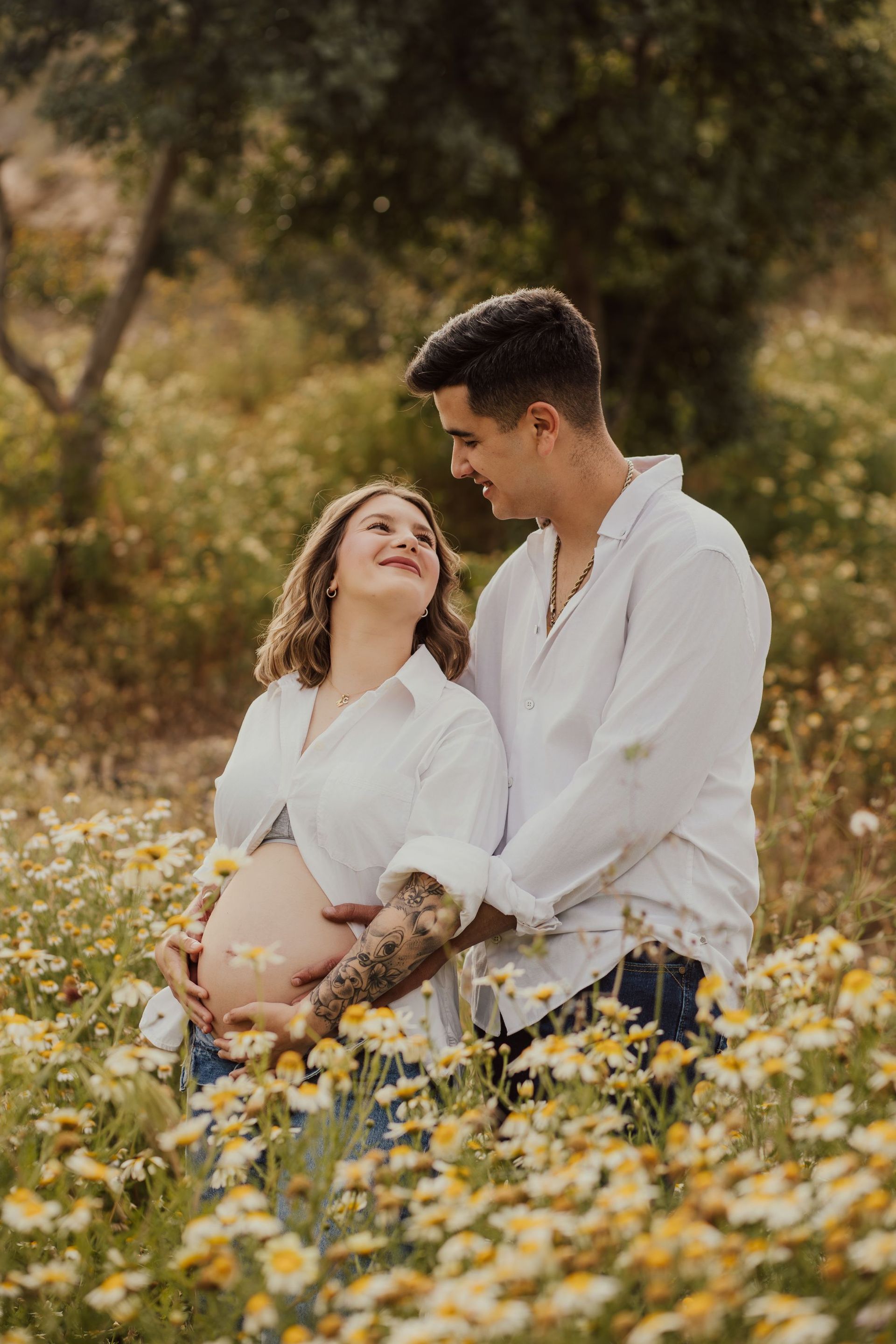 Pareja abrazándose en un campo de flores silvestres, vestidos con camisas blancas, con un cálido fondo exterior difuminado.