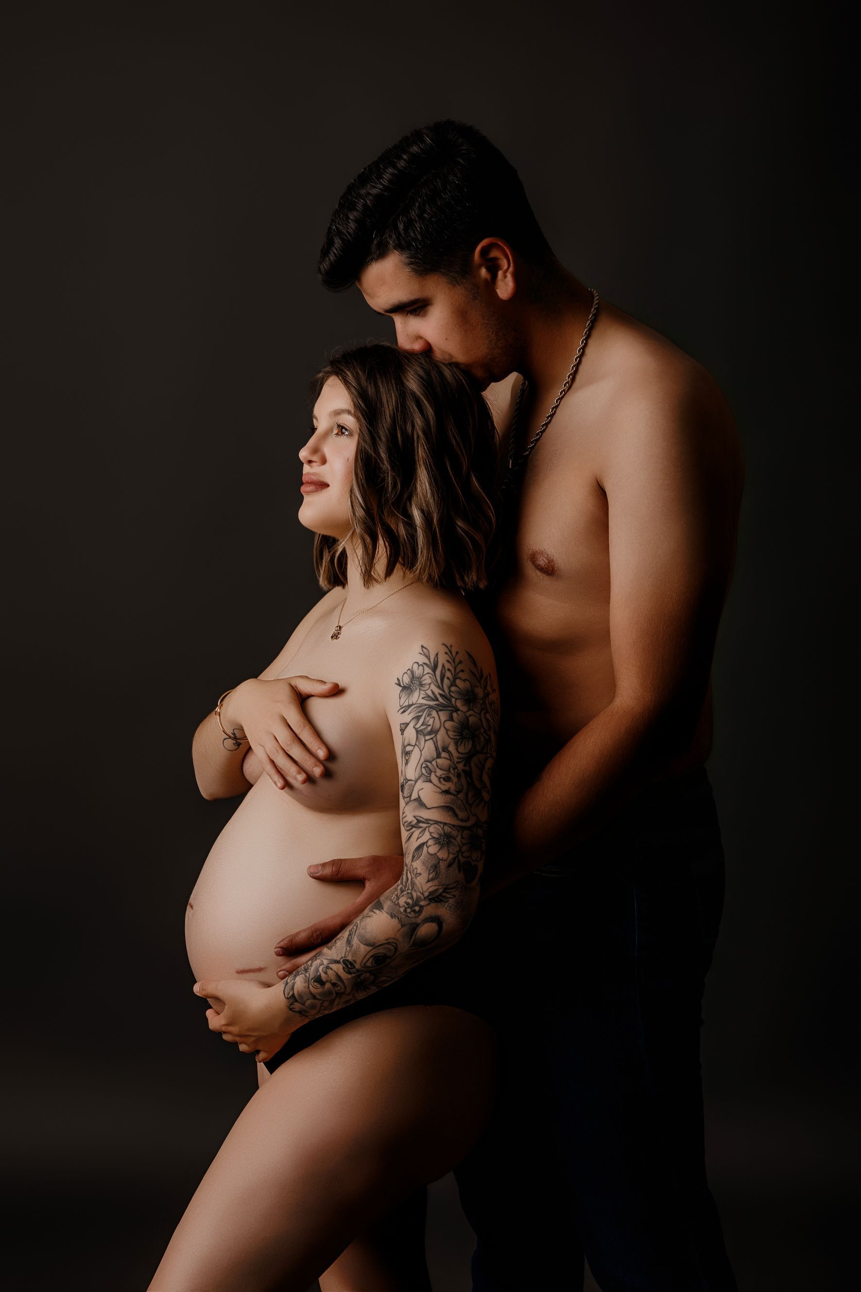 Una mujer embarazada con el brazo tatuado aparece de perfil, abrazada por su pareja sin camisa, sobre un fondo oscuro.