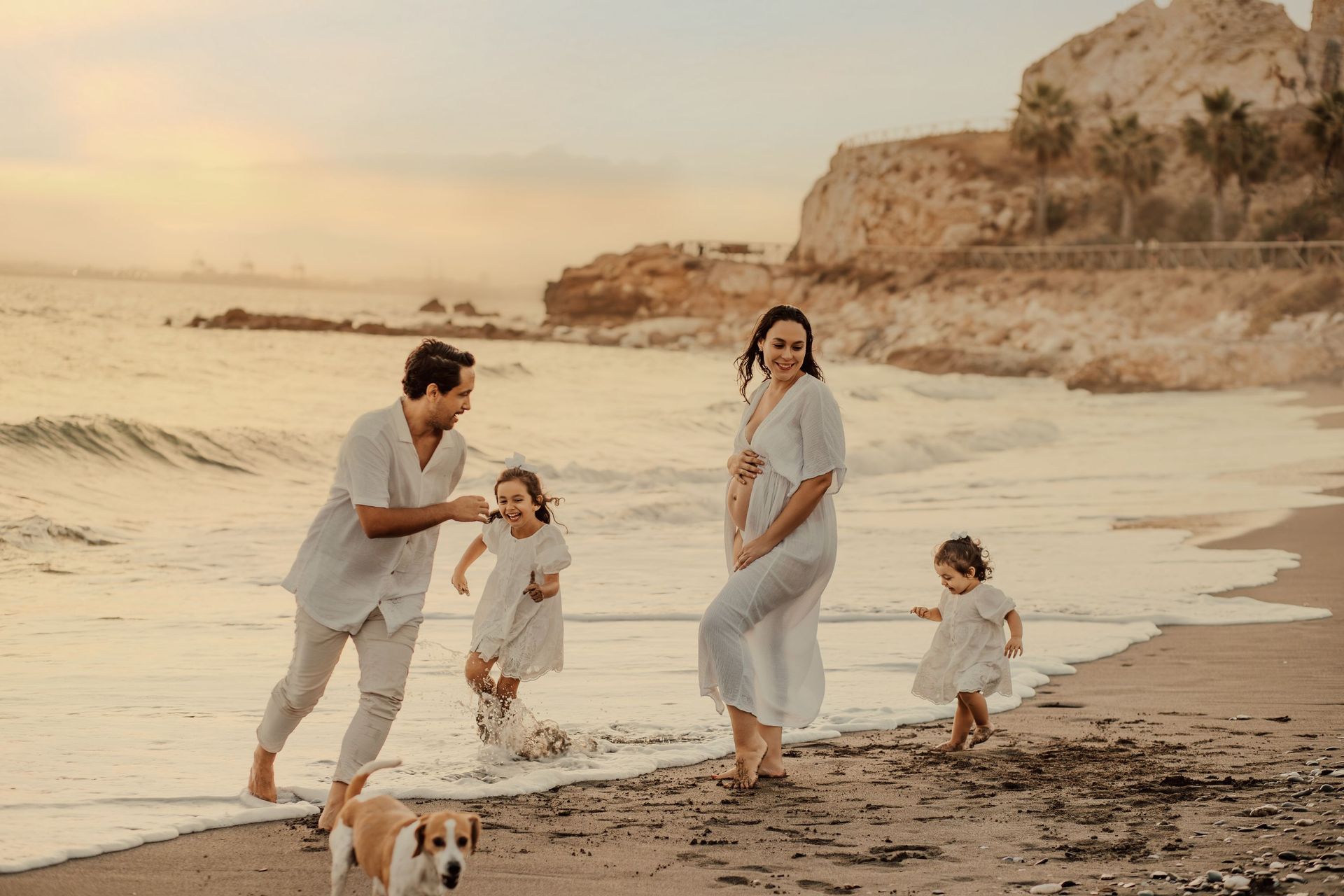 Una familia pasea por la playa al atardecer, con olas, acantilados y un perrito cerca de la orilla.