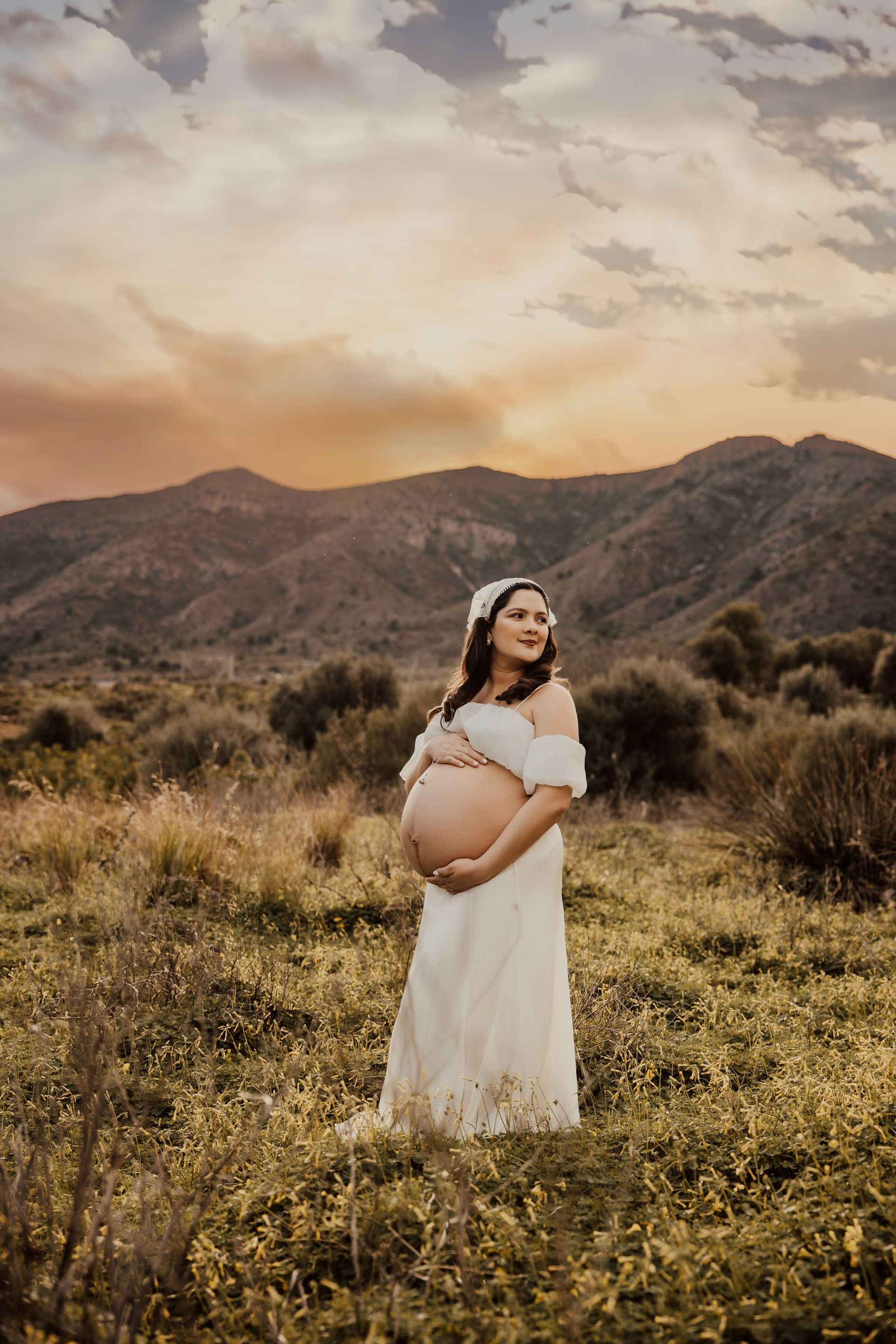Mujer embarazada con un vestido blanco de pie en un campo de hierba al atardecer, con montañas al fondo.