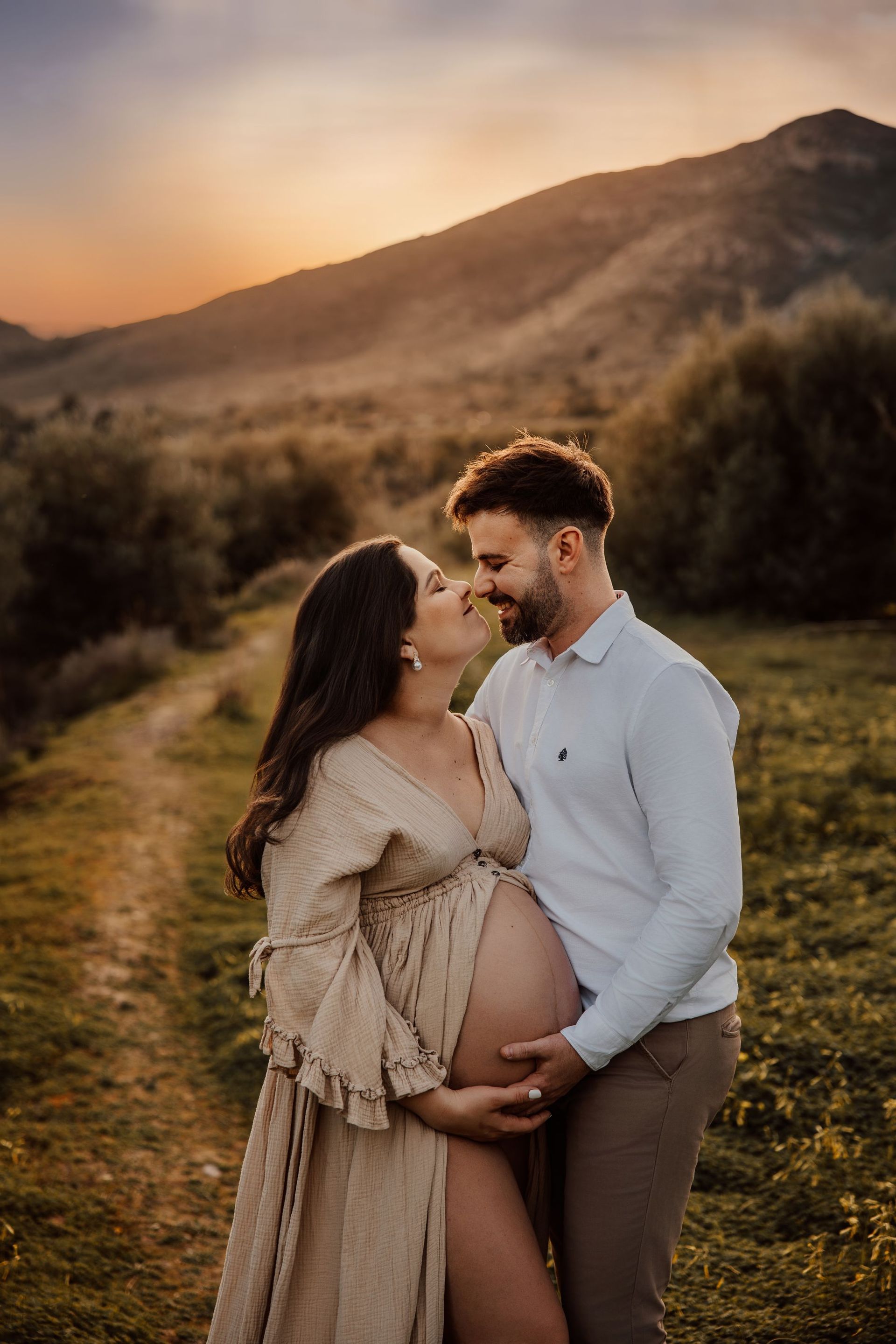 Una pareja embarazada se abraza en un campo al atardecer, con colinas y una cálida luz dorada de fondo.