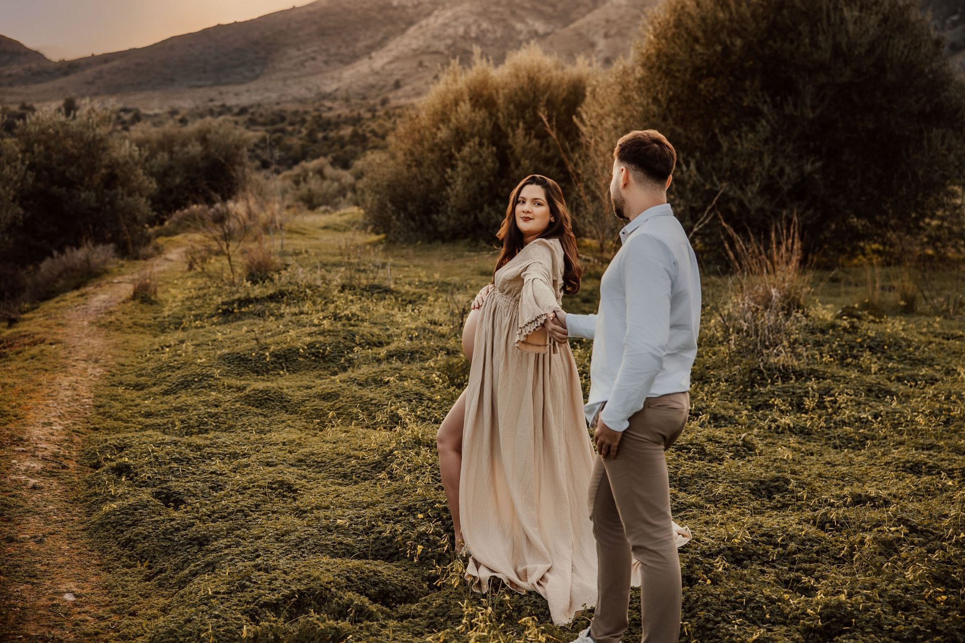 Una pareja de pie en un campo de hierba al atardecer, con colinas y árboles al fondo.